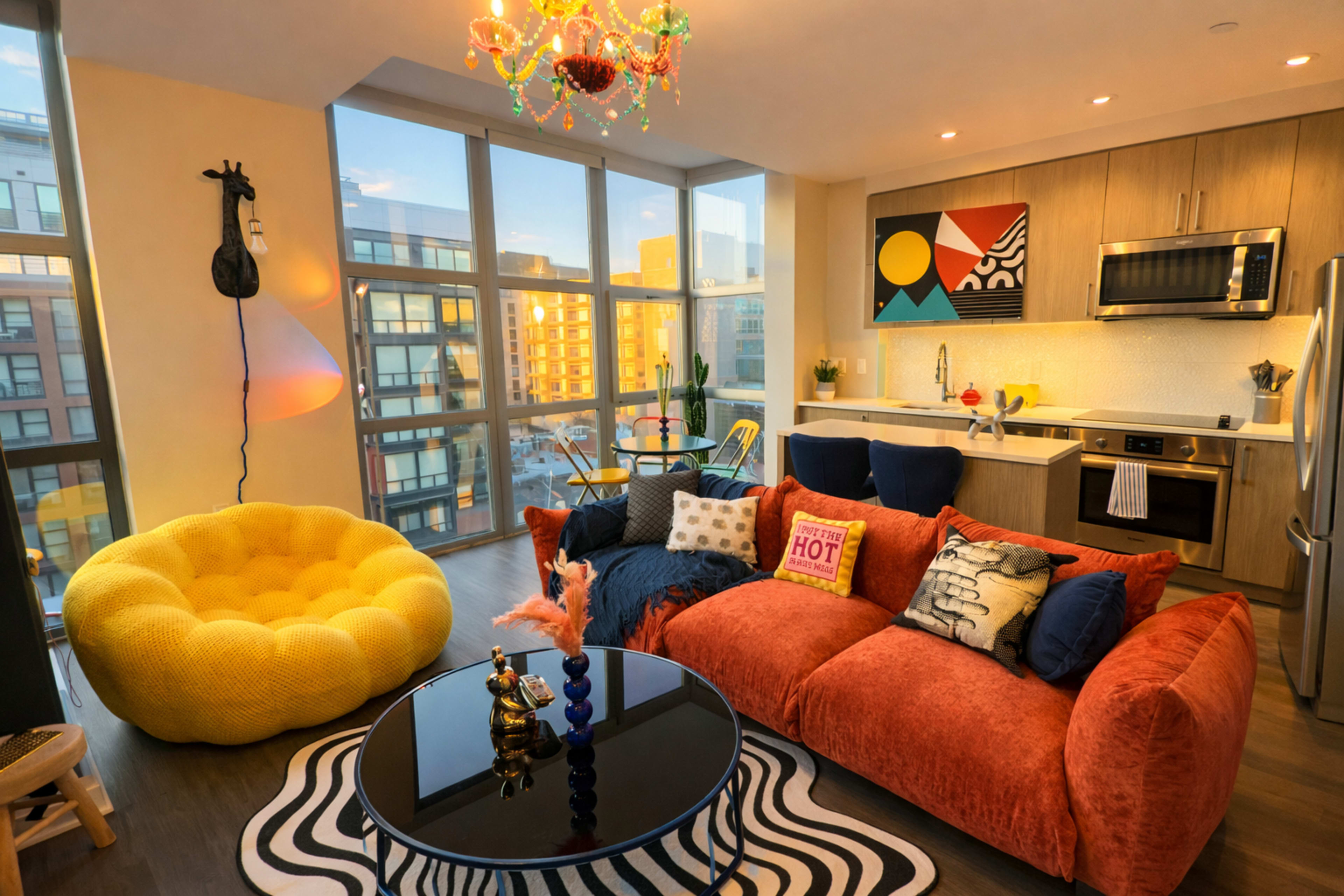 The image depicts a modern living room with a large yellow bean bag, a vibrant orange sofa, a black coffee table, and a kitchen area in the background, all illuminated by natural light from large windows.