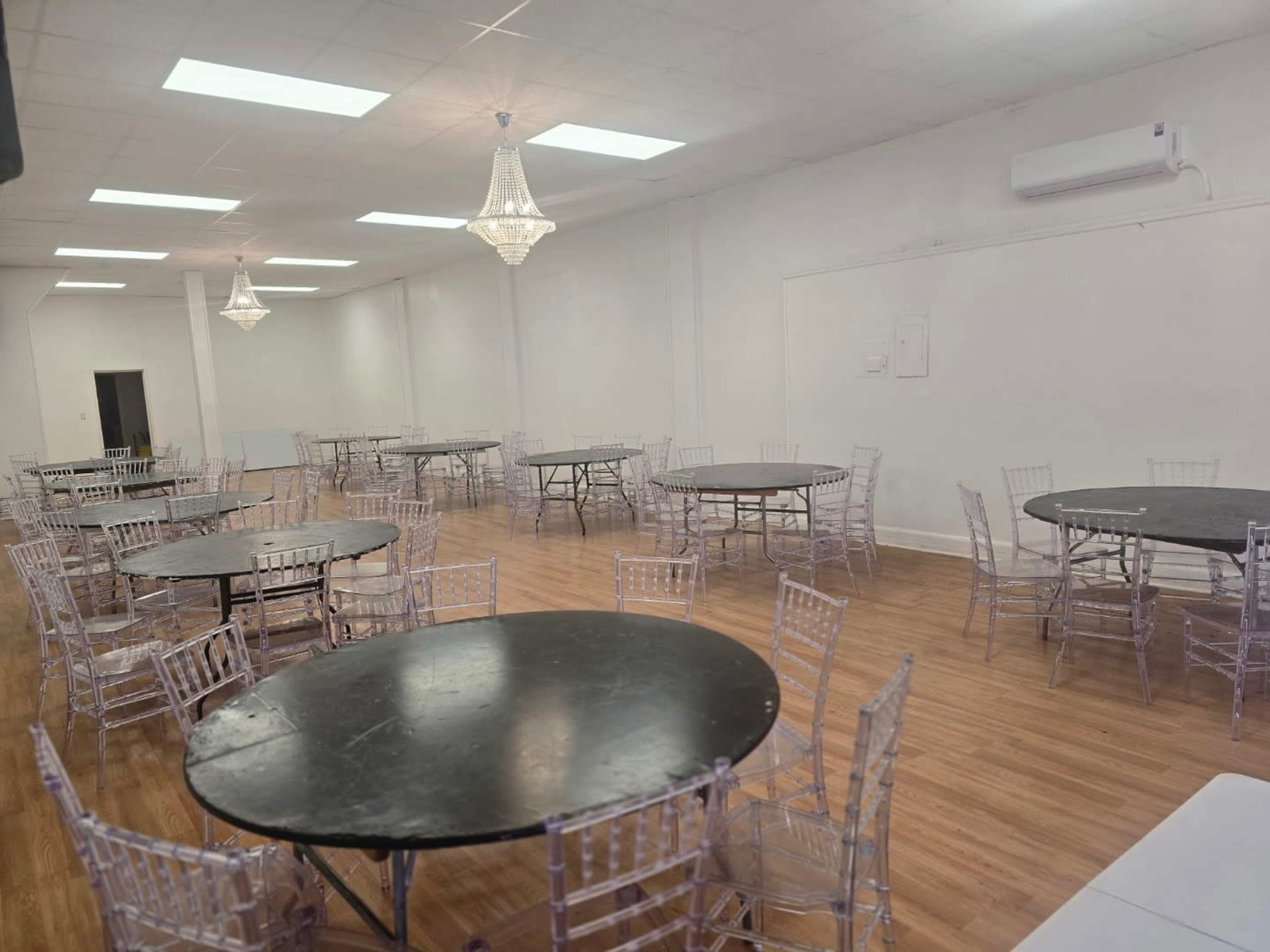 The image shows a spacious event hall with multiple round tables and transparent chairs arranged neatly on a wooden floor, illuminated by overhead lighting and chandeliers.