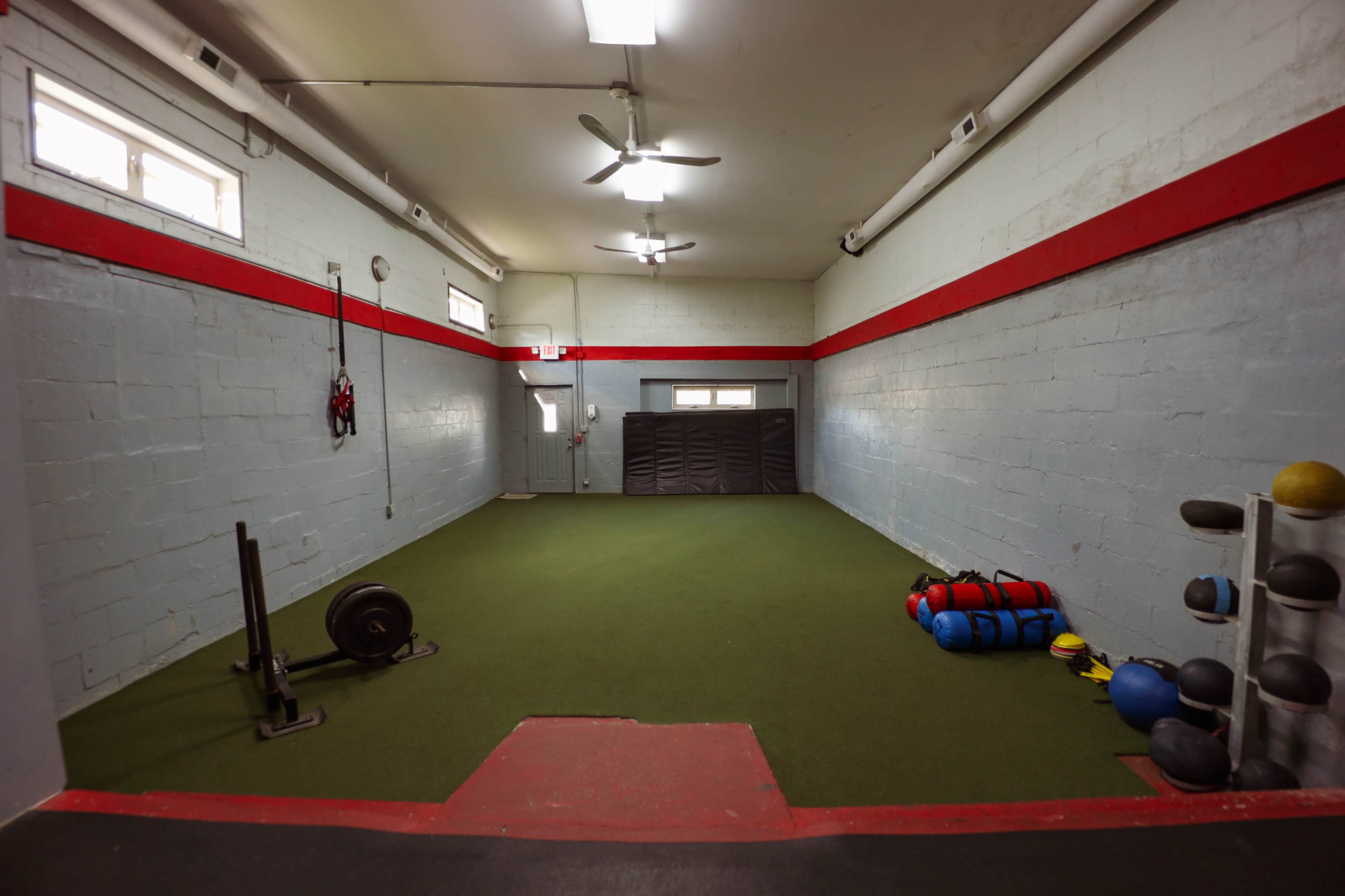 An indoor gym area featuring green turf flooring, red and gray walls, exercise equipment, and wall-mounted fans.