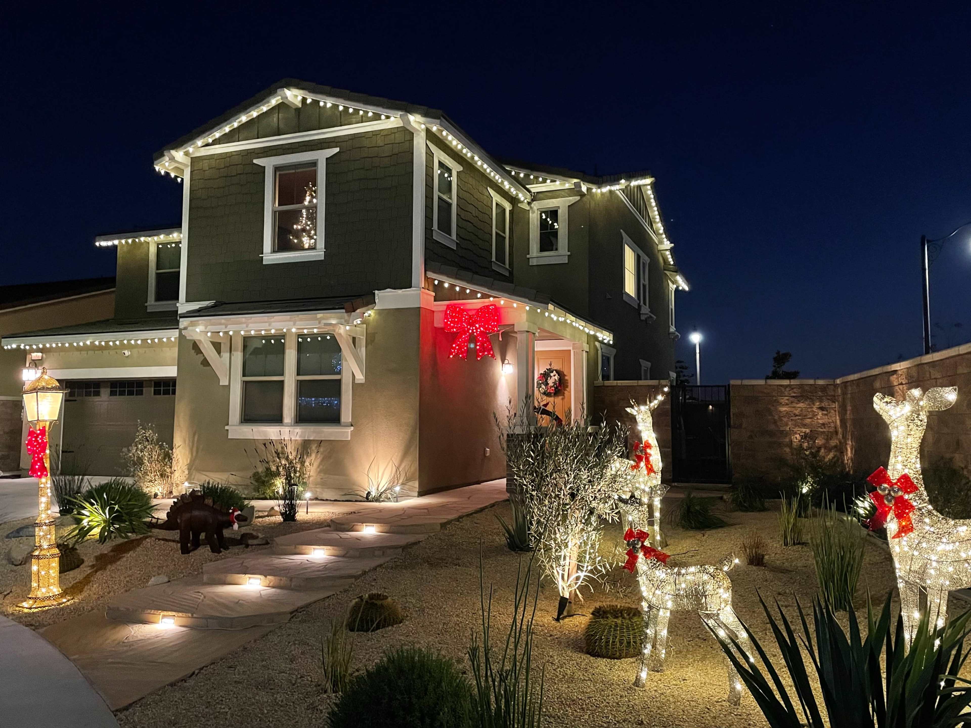 A two-story house is adorned with Christmas lights and decorations, featuring illuminated reindeer in the front yard and a warmly lit pathway.