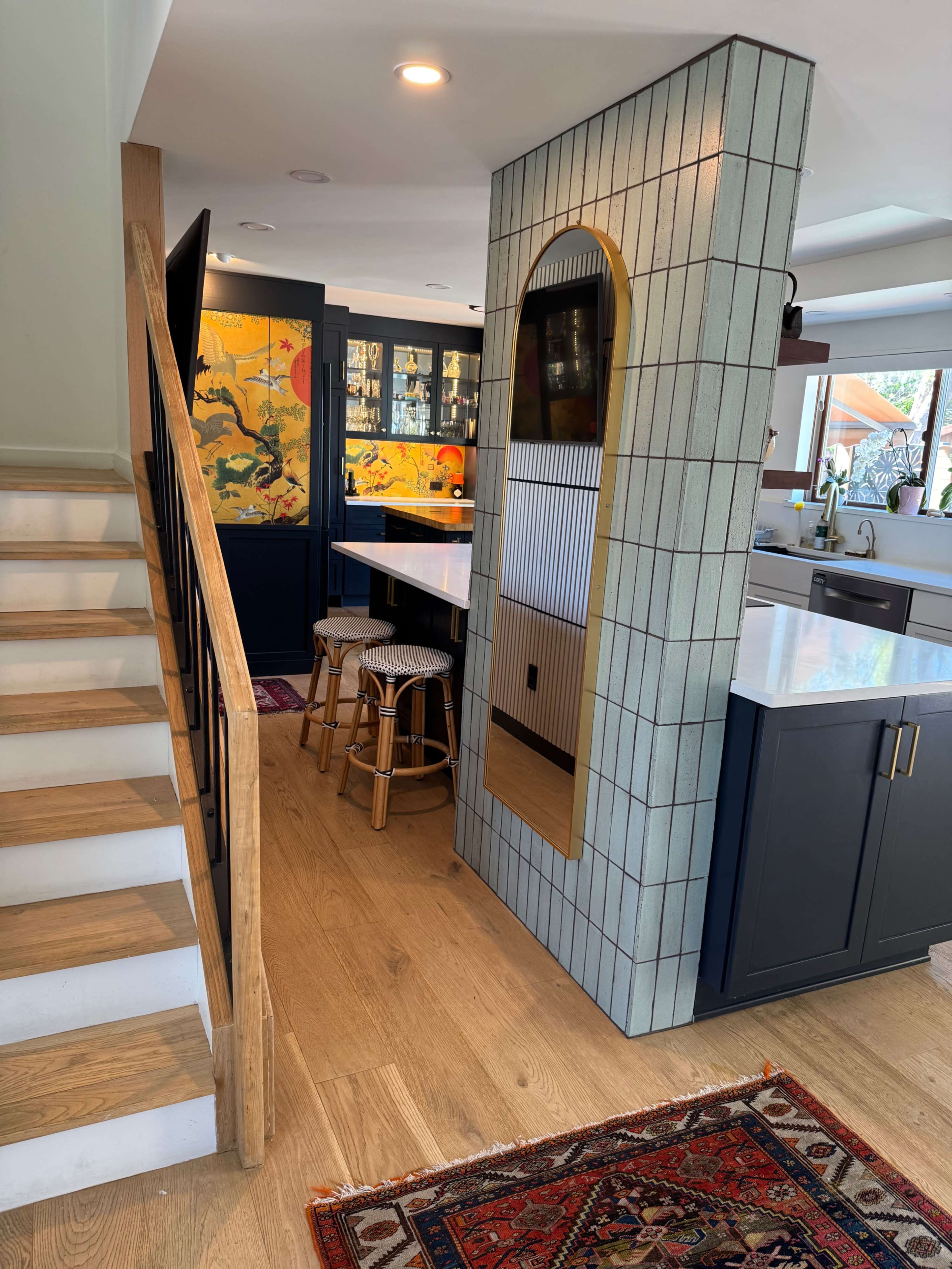 The image shows a modern kitchen area with a staircase on the left, featuring a tiled column, a bar with stools, and a decorative wall with artwork.