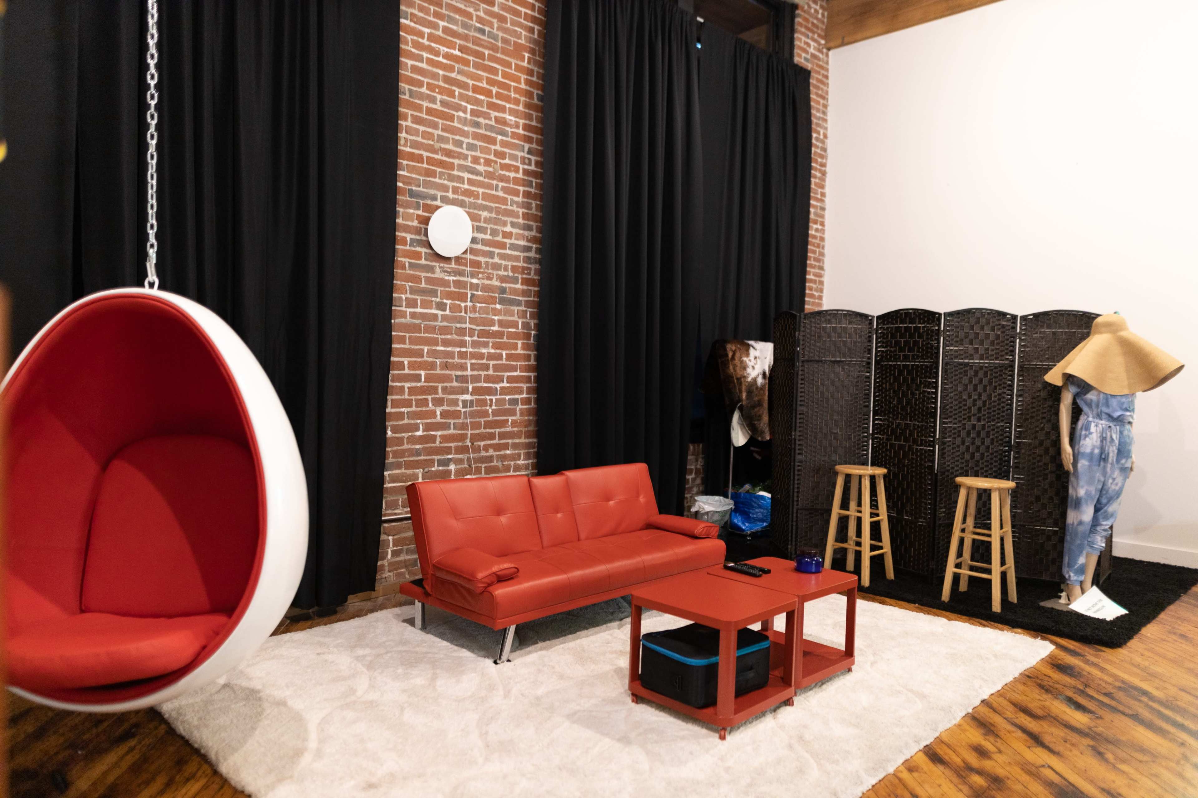 A contemporary living space with a red sofa, a hanging bubble chair, a coffee table, a folding screen, and two wooden stools against a backdrop of exposed brick and black curtains.