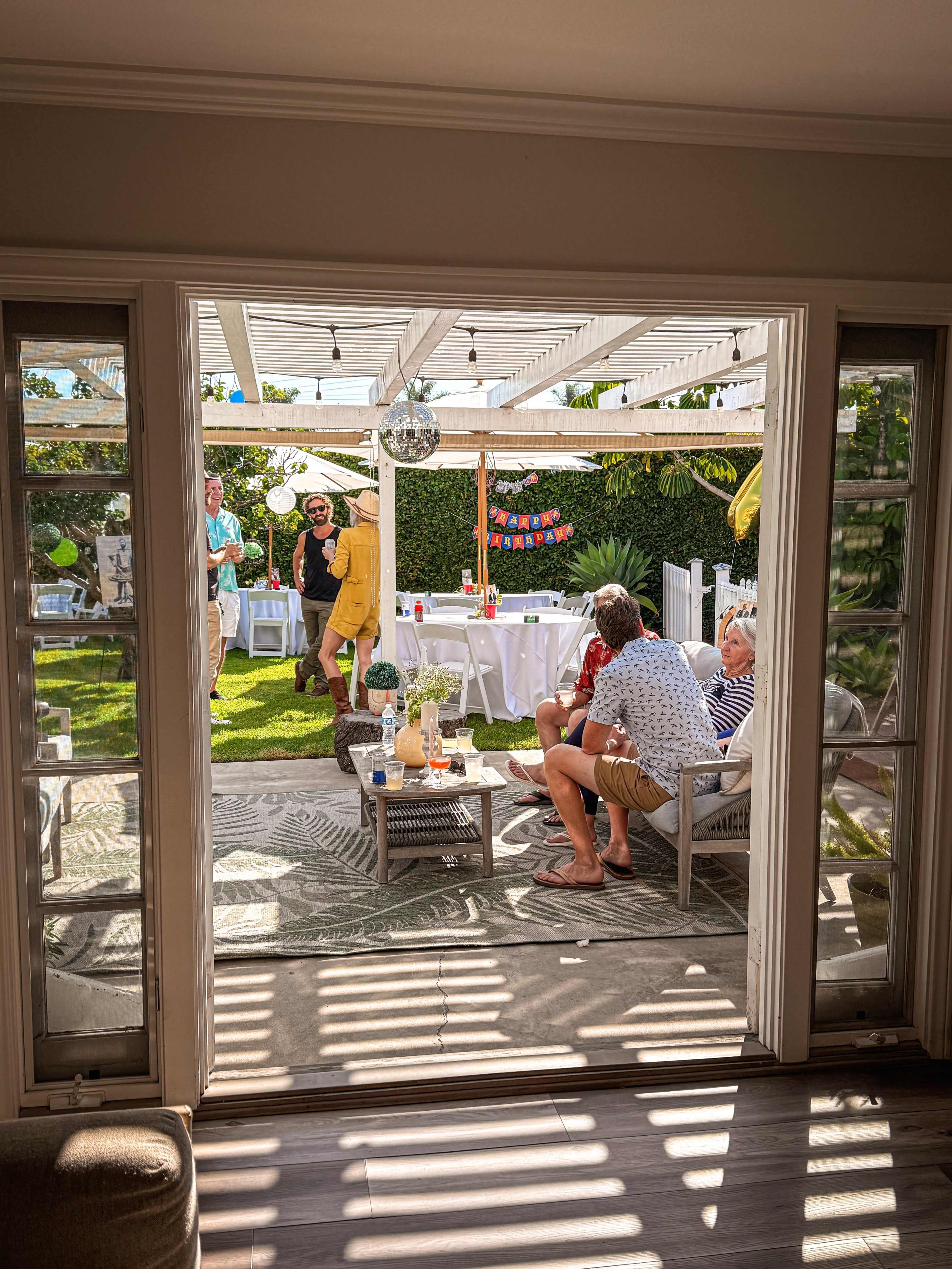 A gathering is in progress in a backyard, with people socializing under a pergola and drinks on a table.