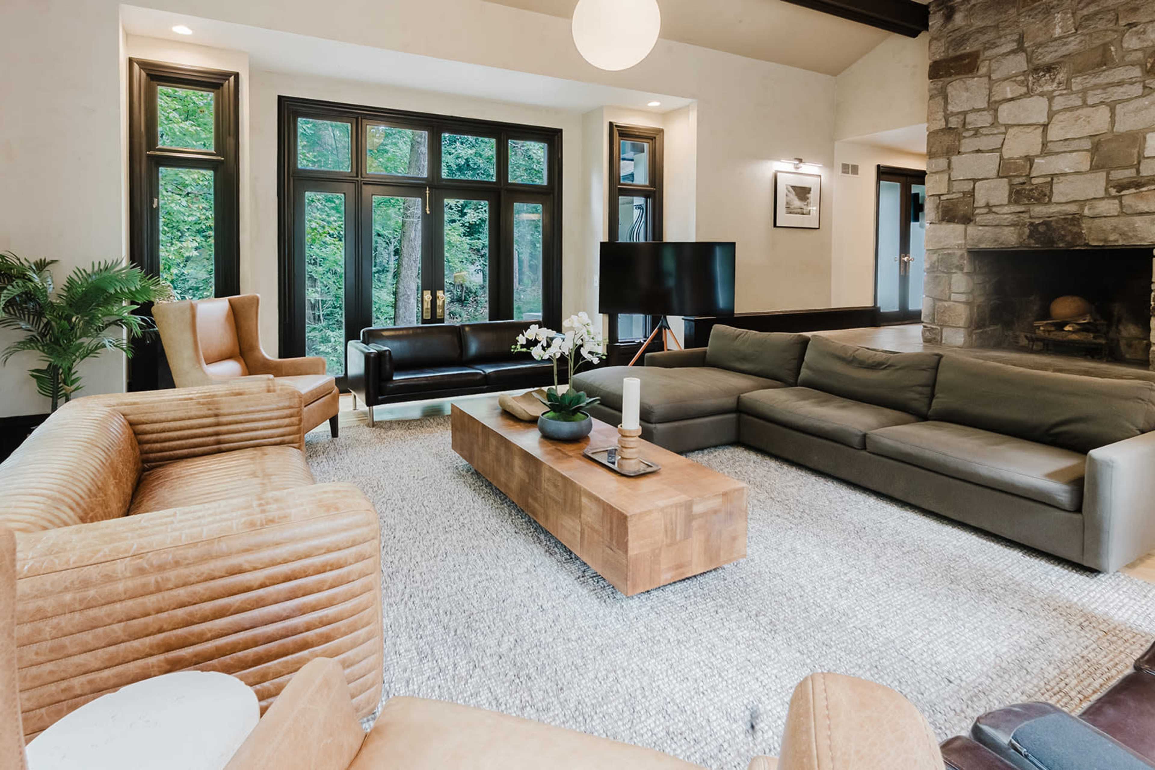 The living room features a mix of modern and traditional furnishings, with a large sectional sofa, a wooden coffee table, and large windows overlooking a natural landscape.
