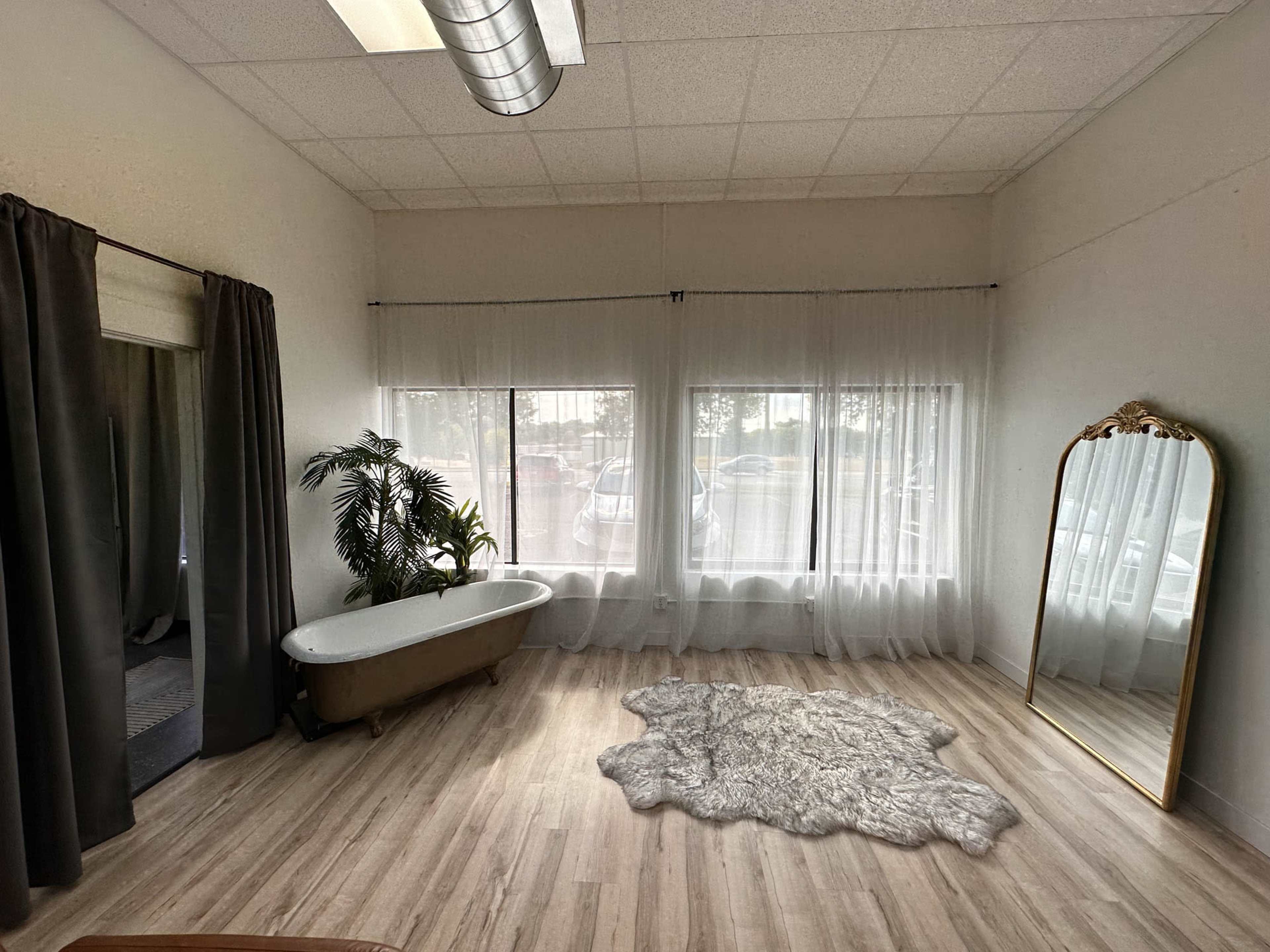 A spacious room featuring large windows, a free-standing bathtub, a decorative mirror, and a textured rug on a wooden floor.