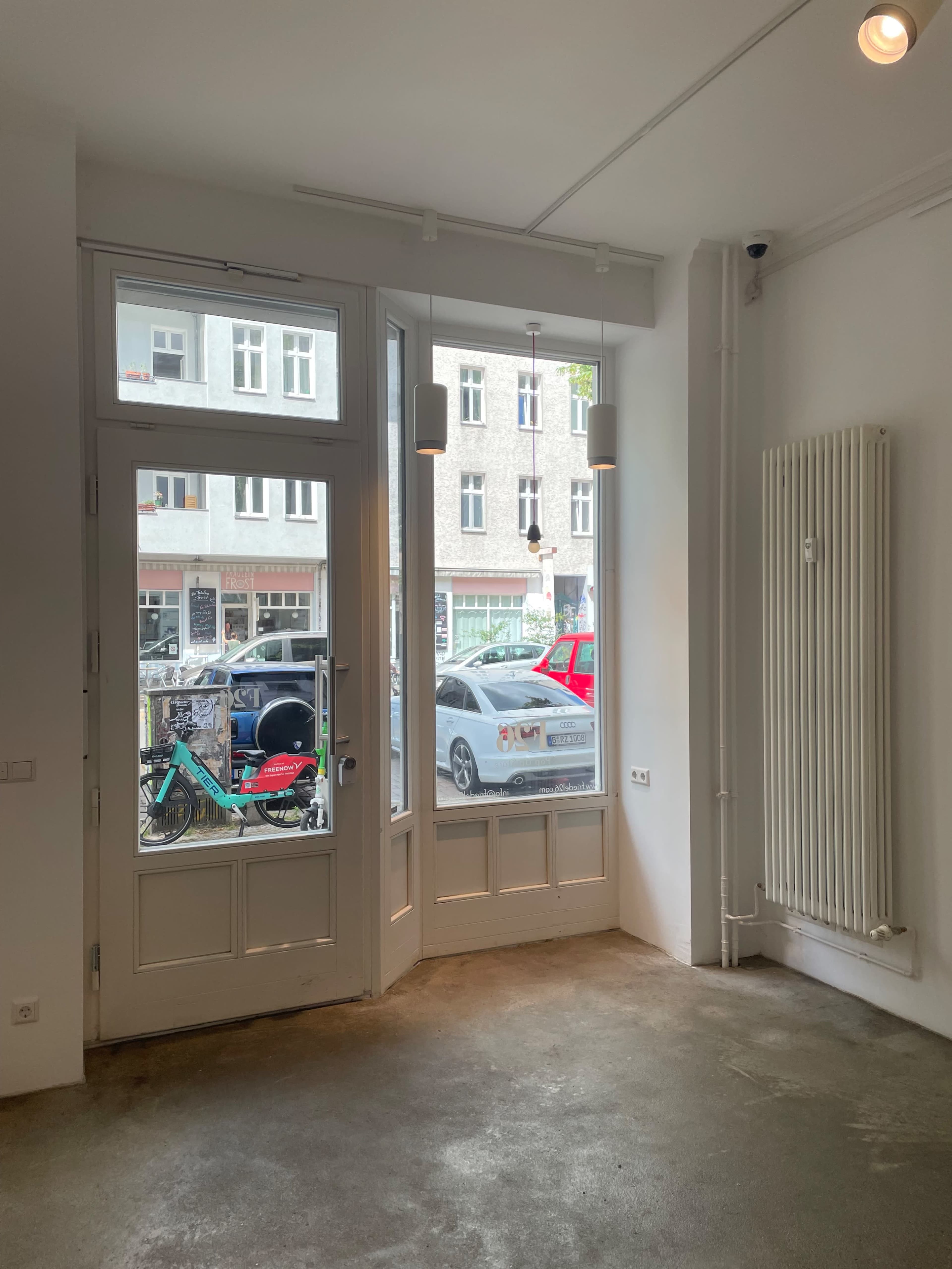 The image shows an empty interior space with large windows facing a street, revealing parked cars and a bicycle outside.