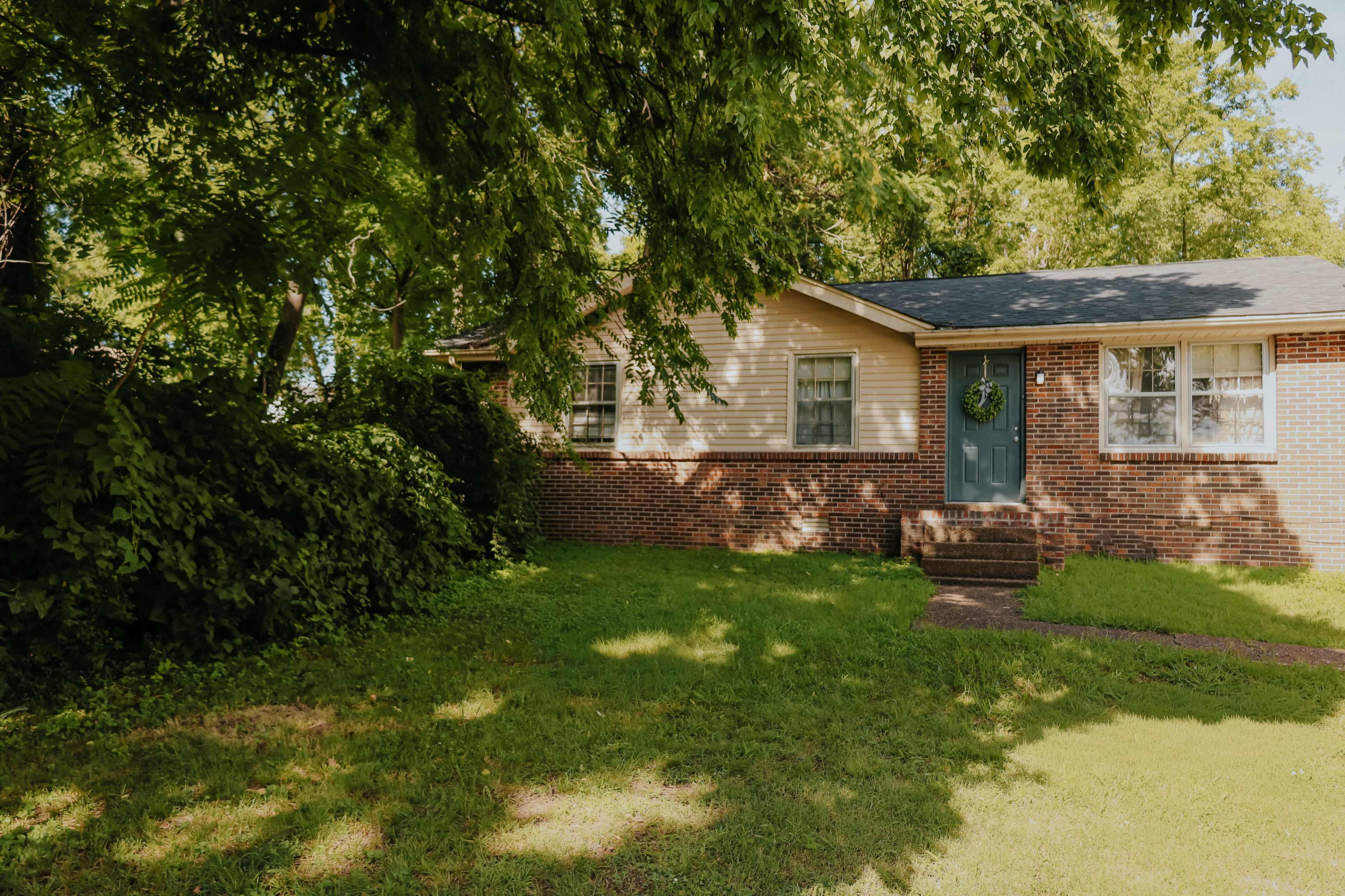 A single-story brick house with a green door and a front porch is surrounded by lush greenery and a well-maintained lawn.