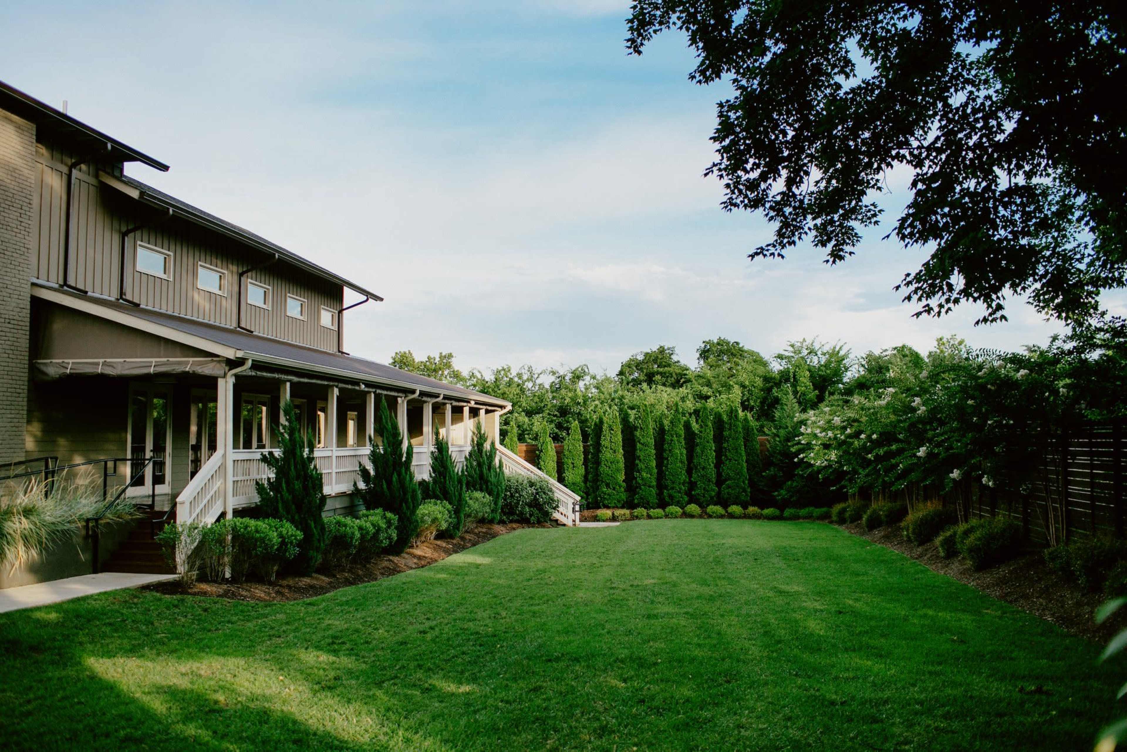 A well-maintained lawn is surrounded by a modern building and tall evergreen hedges under a clear sky.
