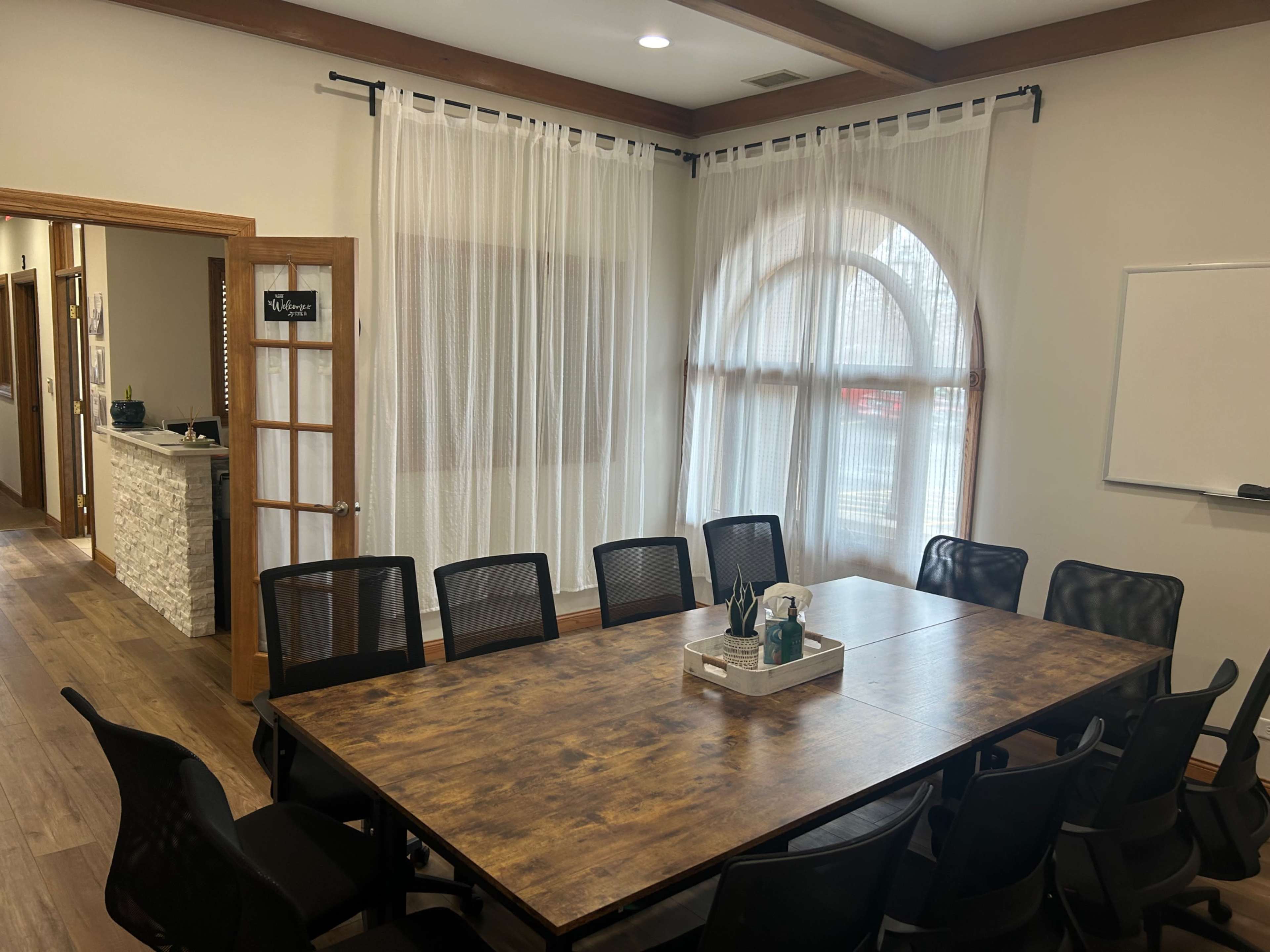 The image shows a conference room with a large wooden table surrounded by black chairs, featuring large windows with sheer curtains and a whiteboard on the wall.