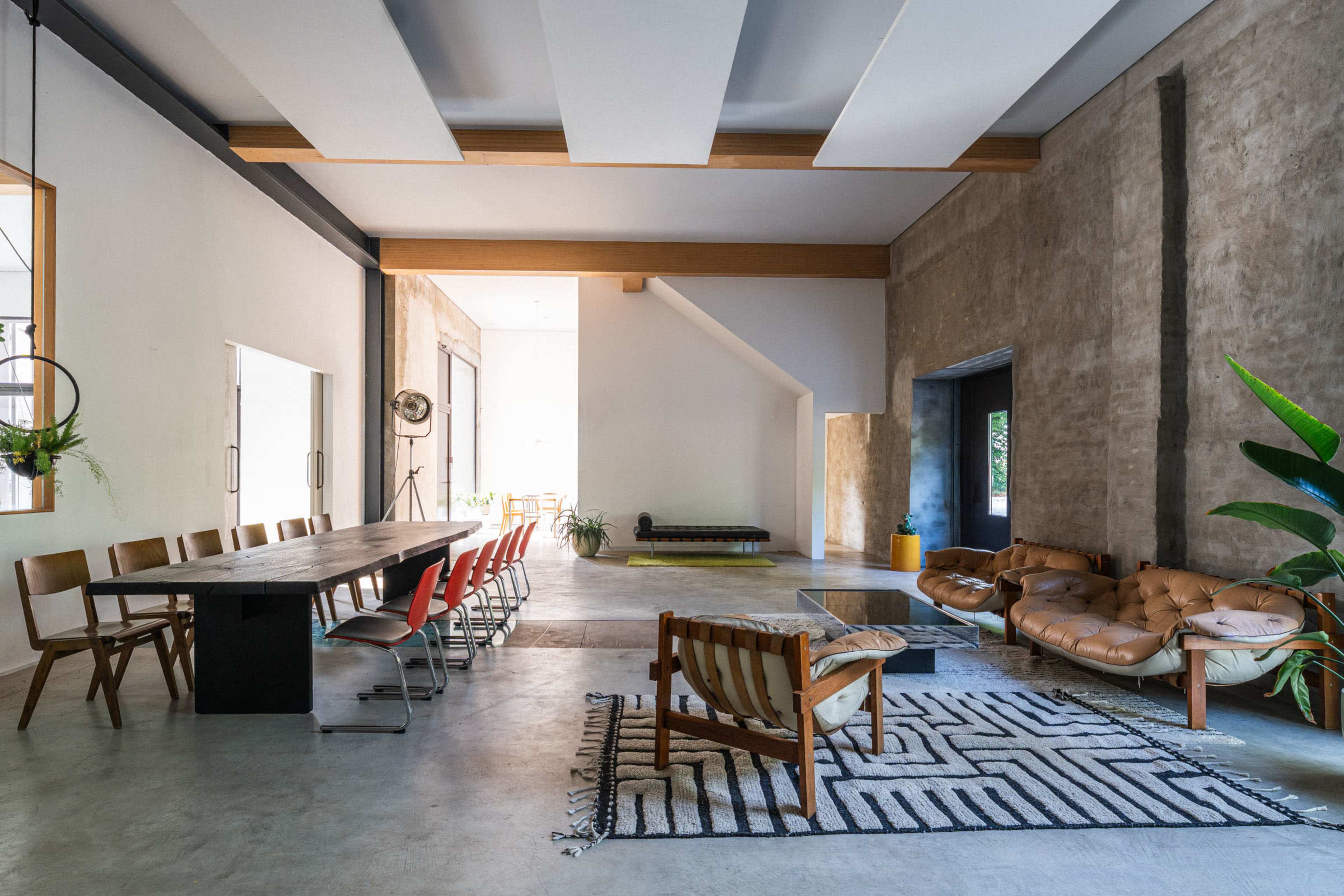 The image shows a spacious, modern interior with a long dining table surrounded by chairs, a seating area with a couch and armchair, and large windows allowing natural light to enter.