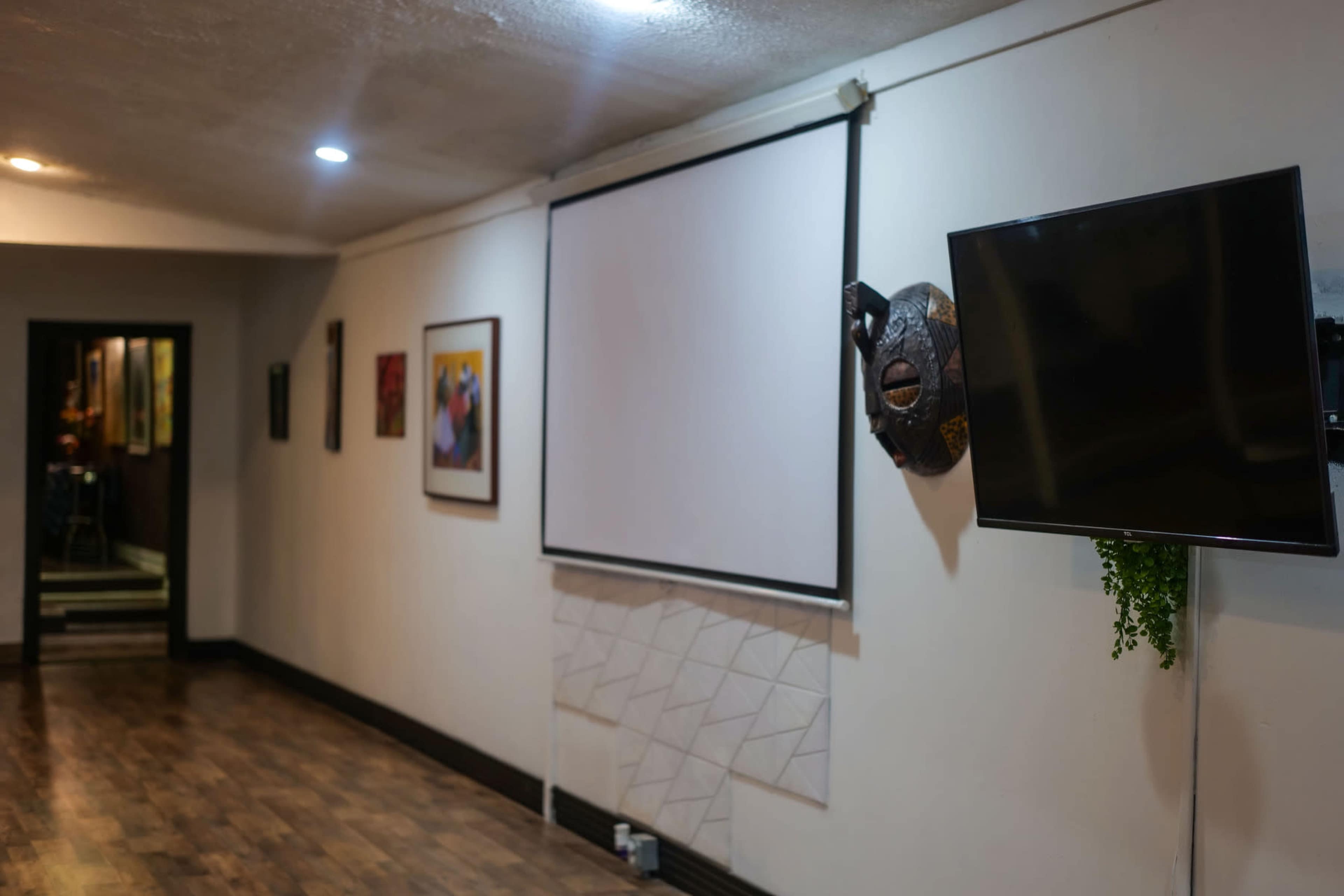 The image shows a room with a blank projection screen mounted on a wall, a flat-screen TV, and framed pictures hanging nearby.