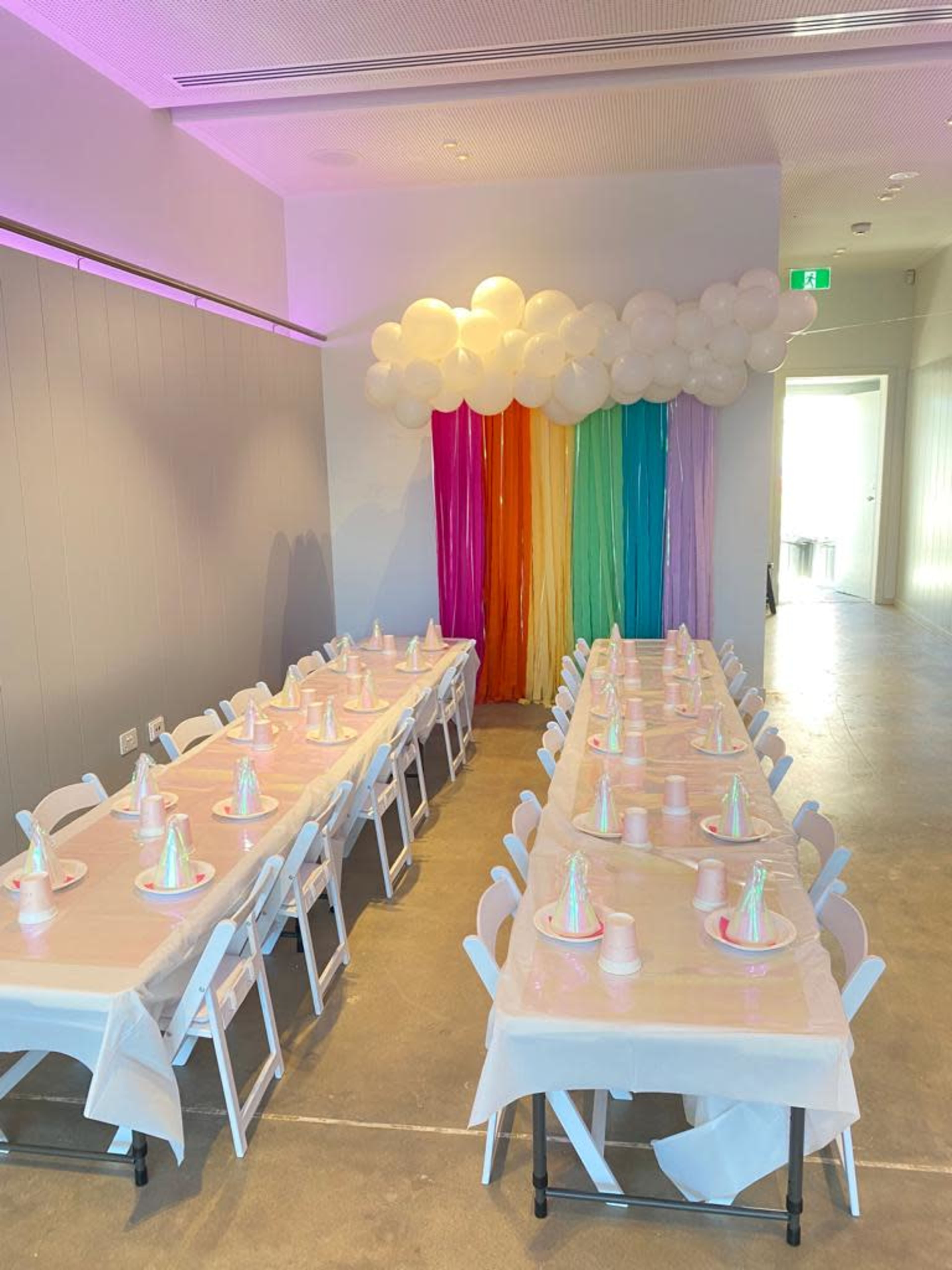 A long, rectangular table is set with pastel-colored party hats and plates beneath a rainbow curtain and decorative balloon cloud.