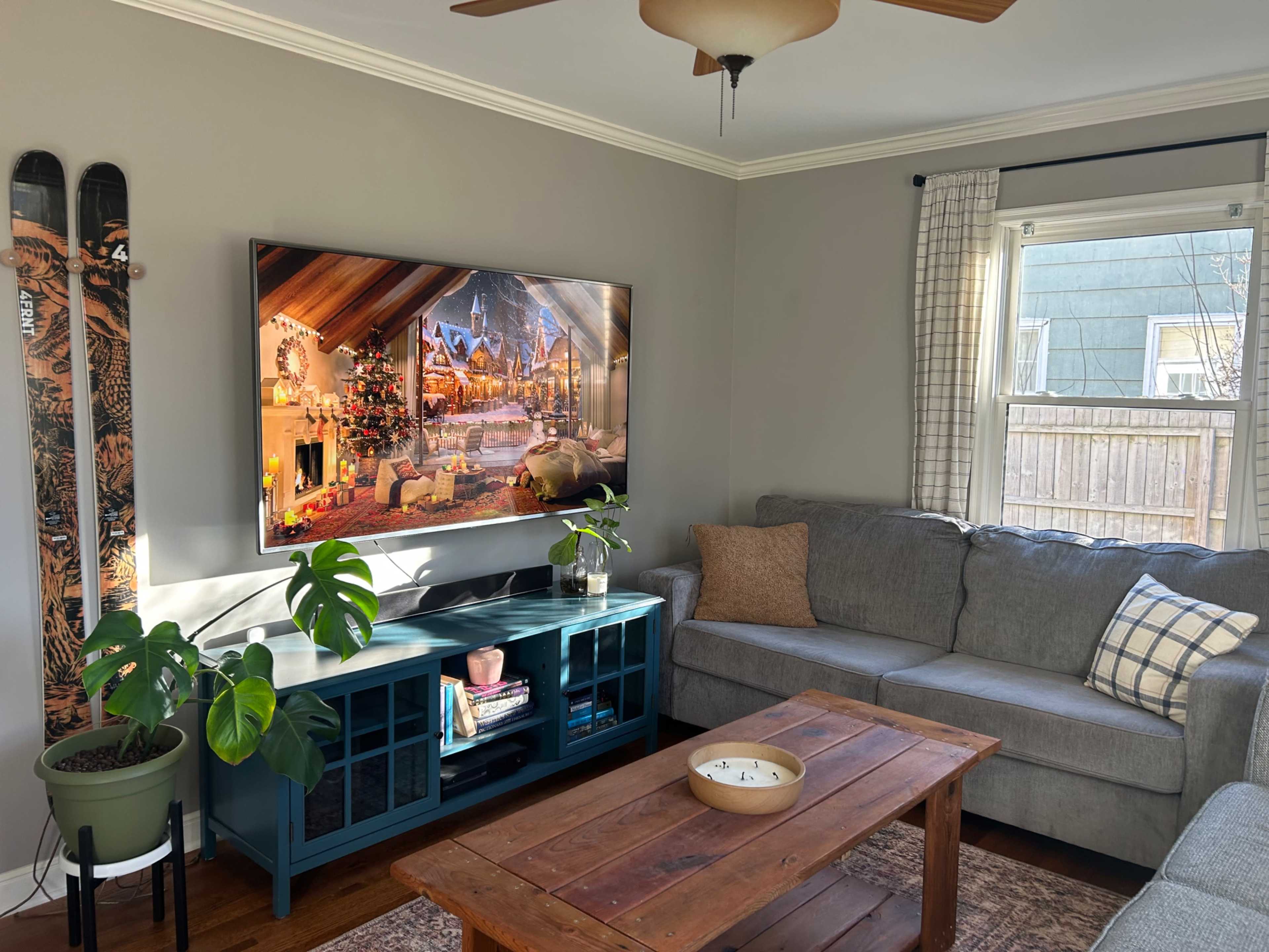 A cozy living room features a gray sofa, a teal coffee table, and a large TV displaying a festive scene, with a potted plant and skis leaning against the wall.