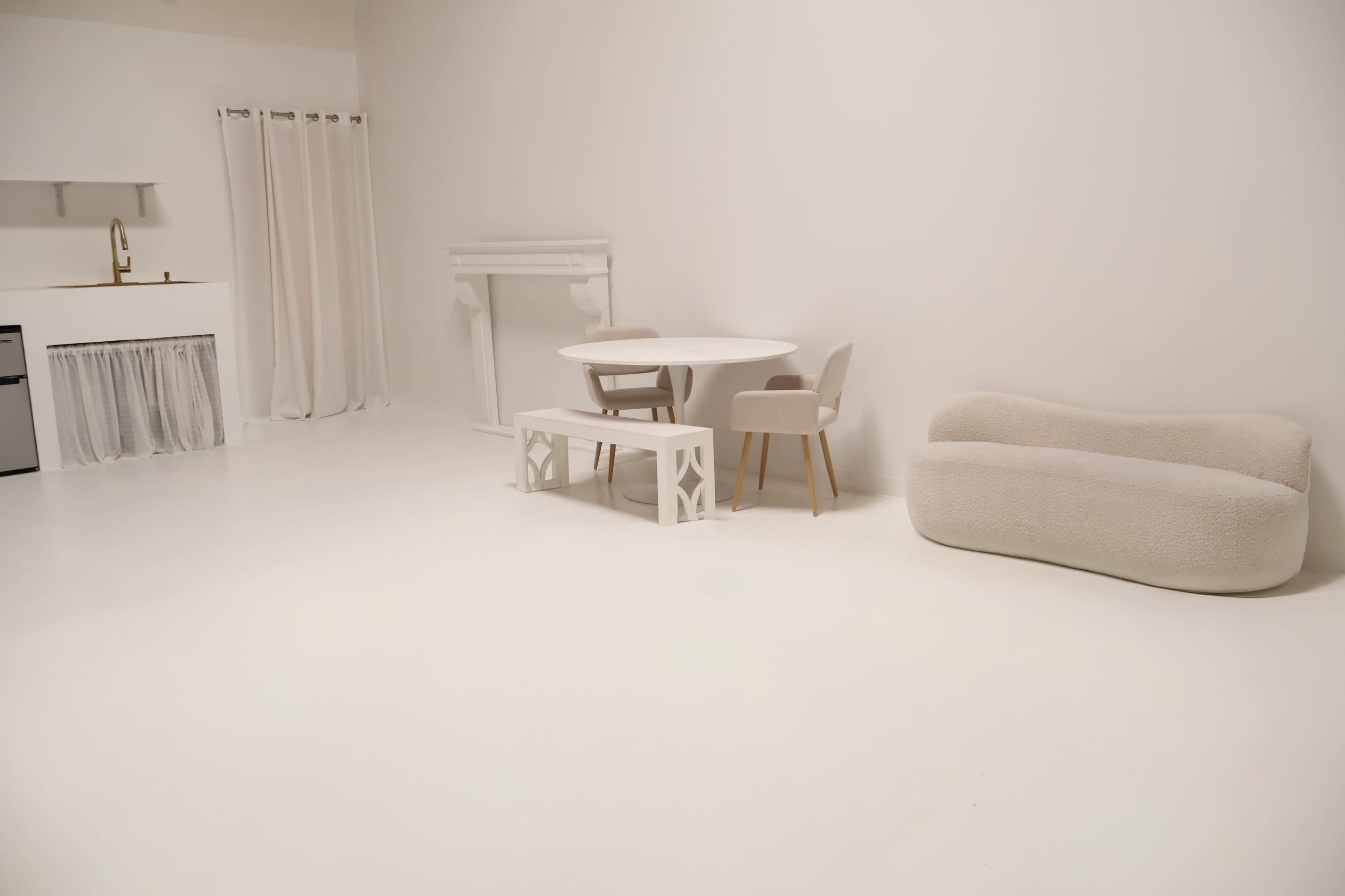 The image shows a minimalist interior space with white walls and flooring, featuring a white table, two chairs, a small bench, and a light-colored sofa.