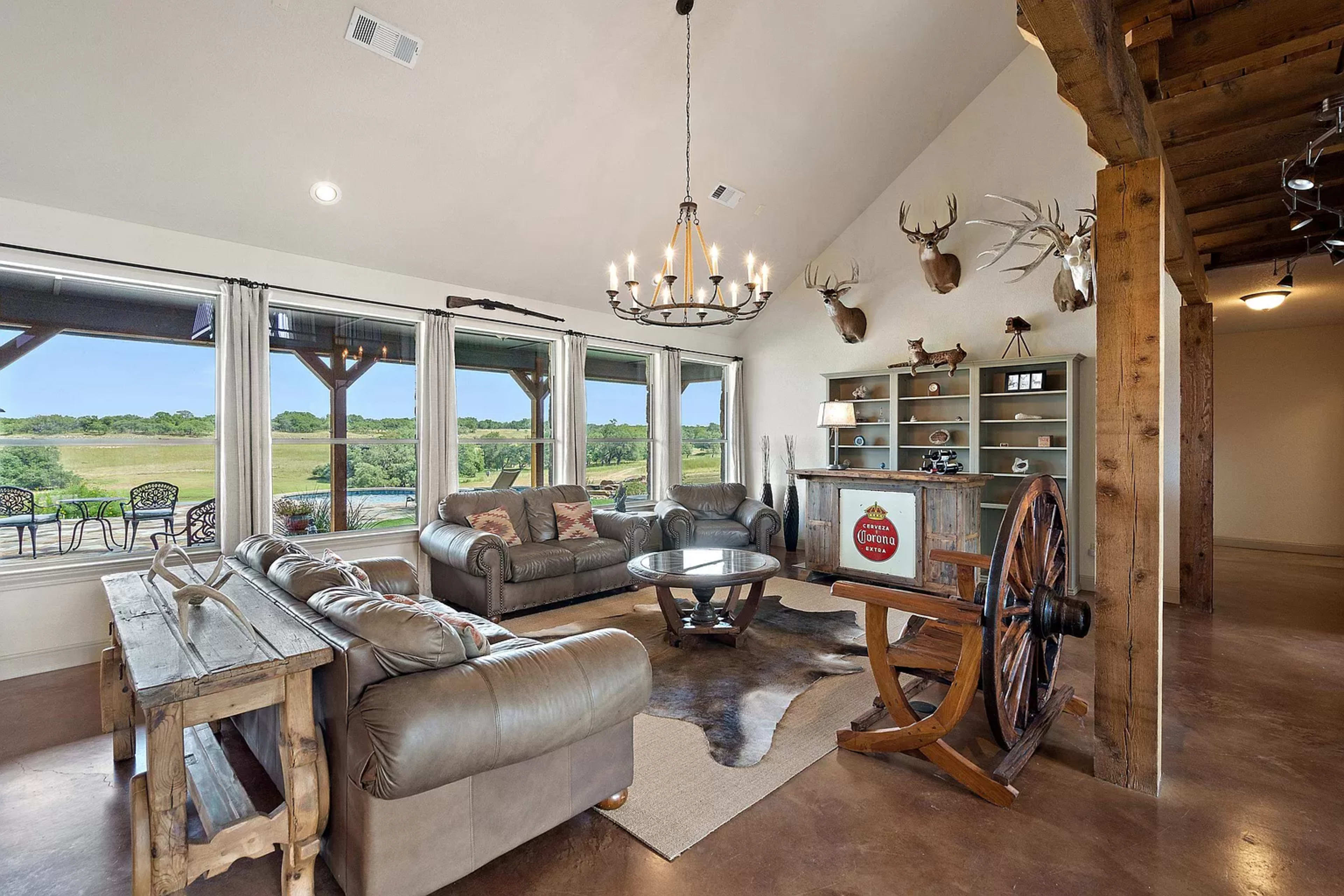 The image shows a spacious living room with large windows overlooking a grassy landscape, featuring a mix of rustic wooden furniture, a cowhide rug, and mounted deer heads on the walls.