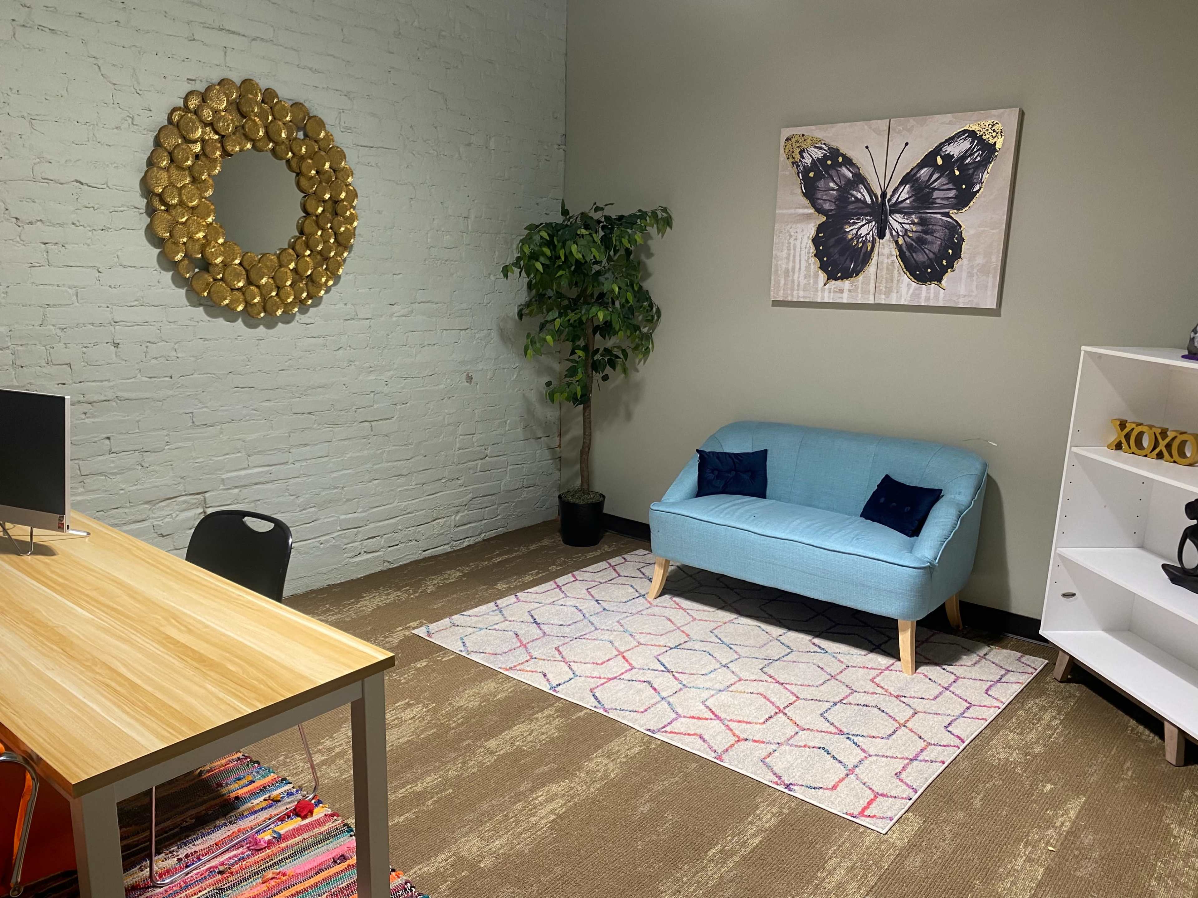 A small office space featuring a blue sofa, a wooden desk with a computer, a decorative butterfly artwork on the wall, and a potted plant.