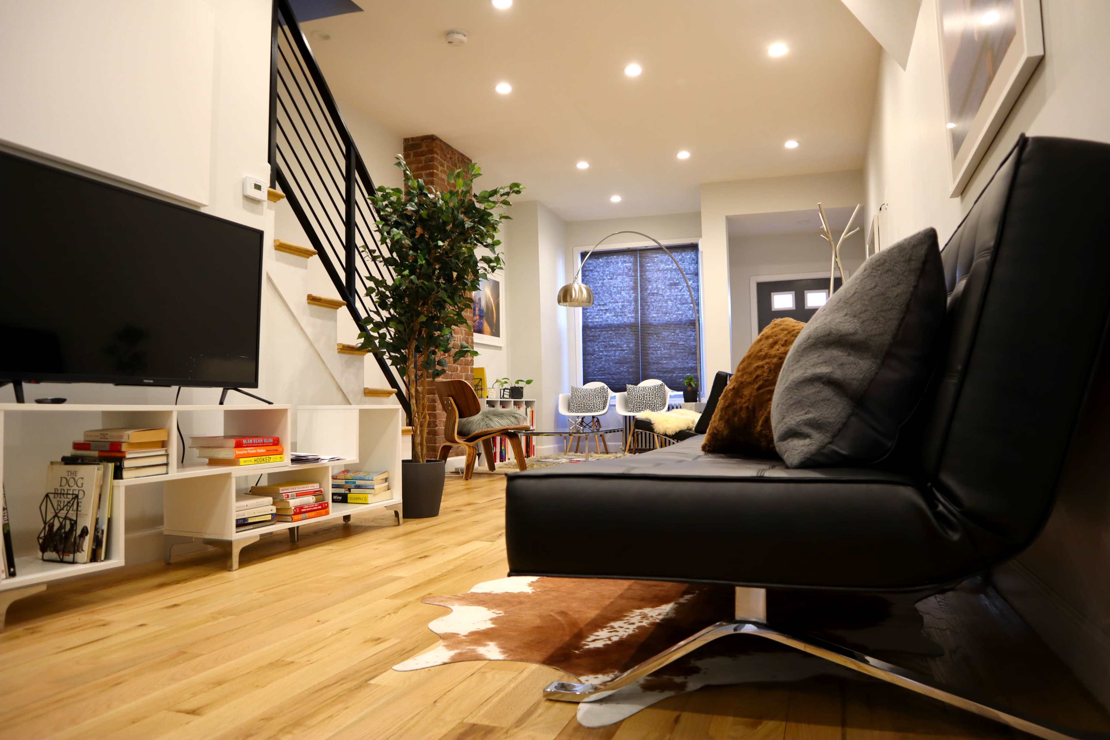 The scene features a modern living room with a black sofa, a television on a white shelf, a potted plant, and a staircase that leads to an upper level.