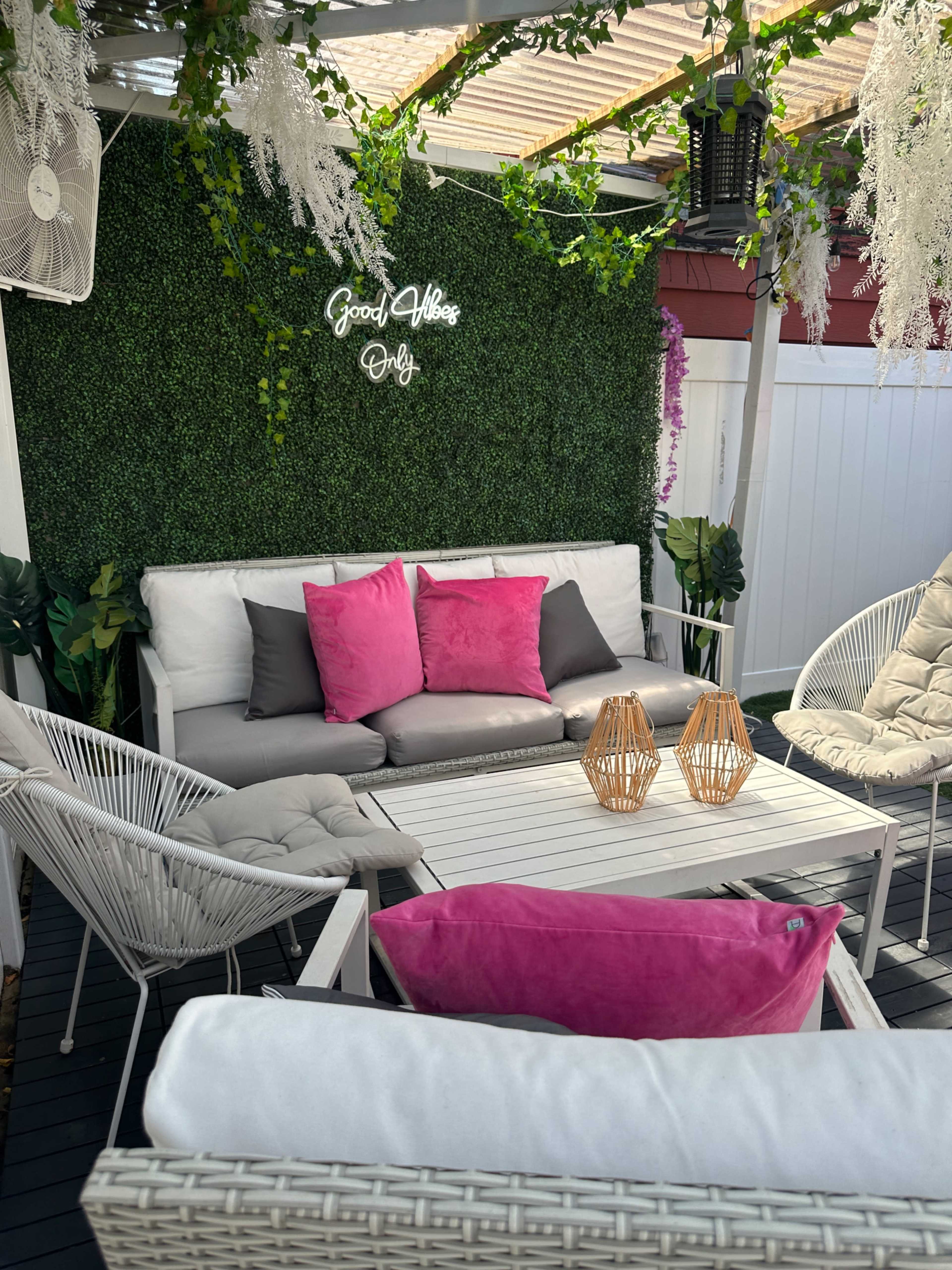 A cozy outdoor seating area with a green wall decor, a white sofa adorned with pink and gray cushions, and decorative wooden lanterns on a white table.