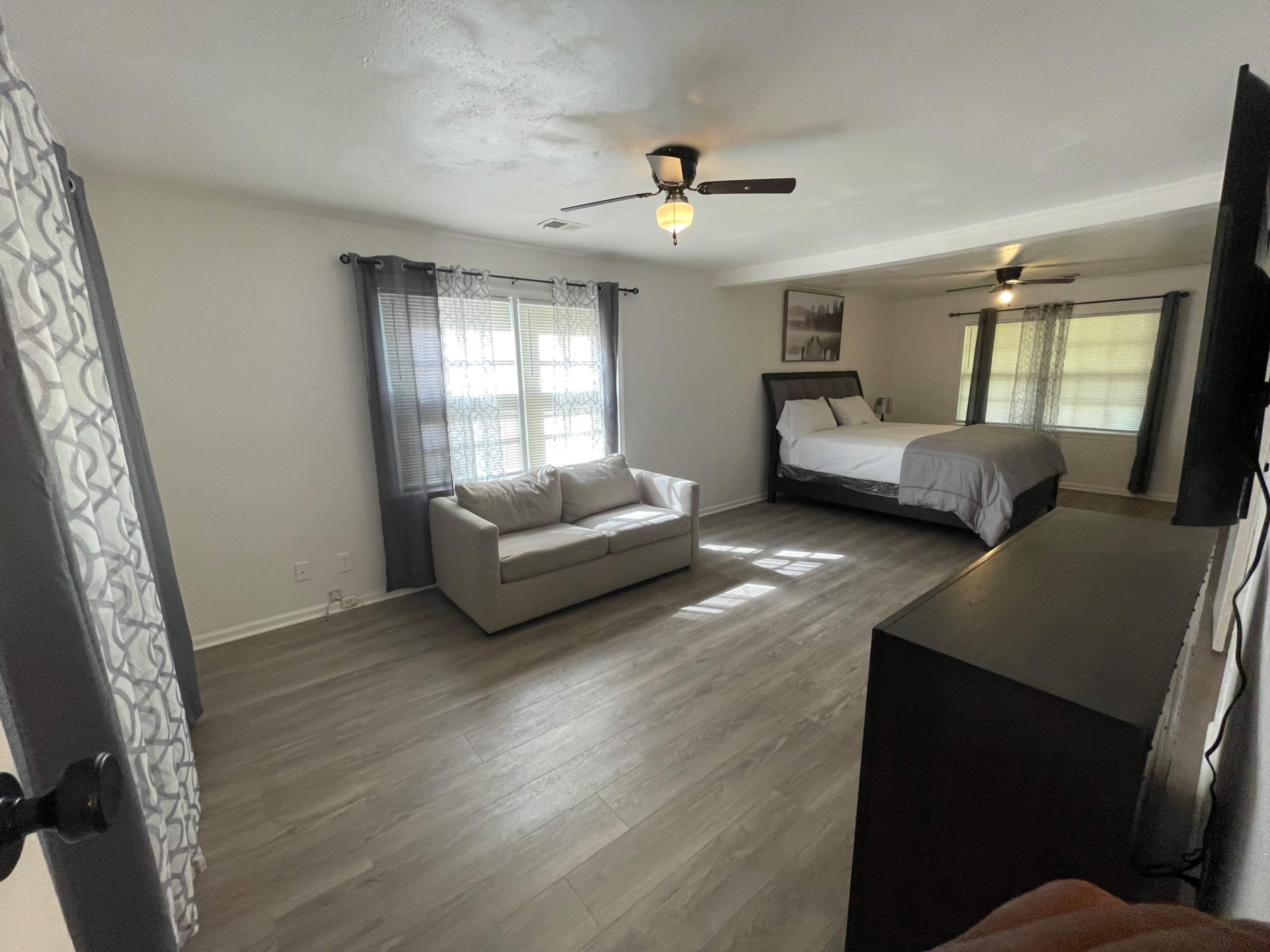 The image shows a spacious bedroom with a queen-sized bed, a small couch, and large windows adorned with curtains.