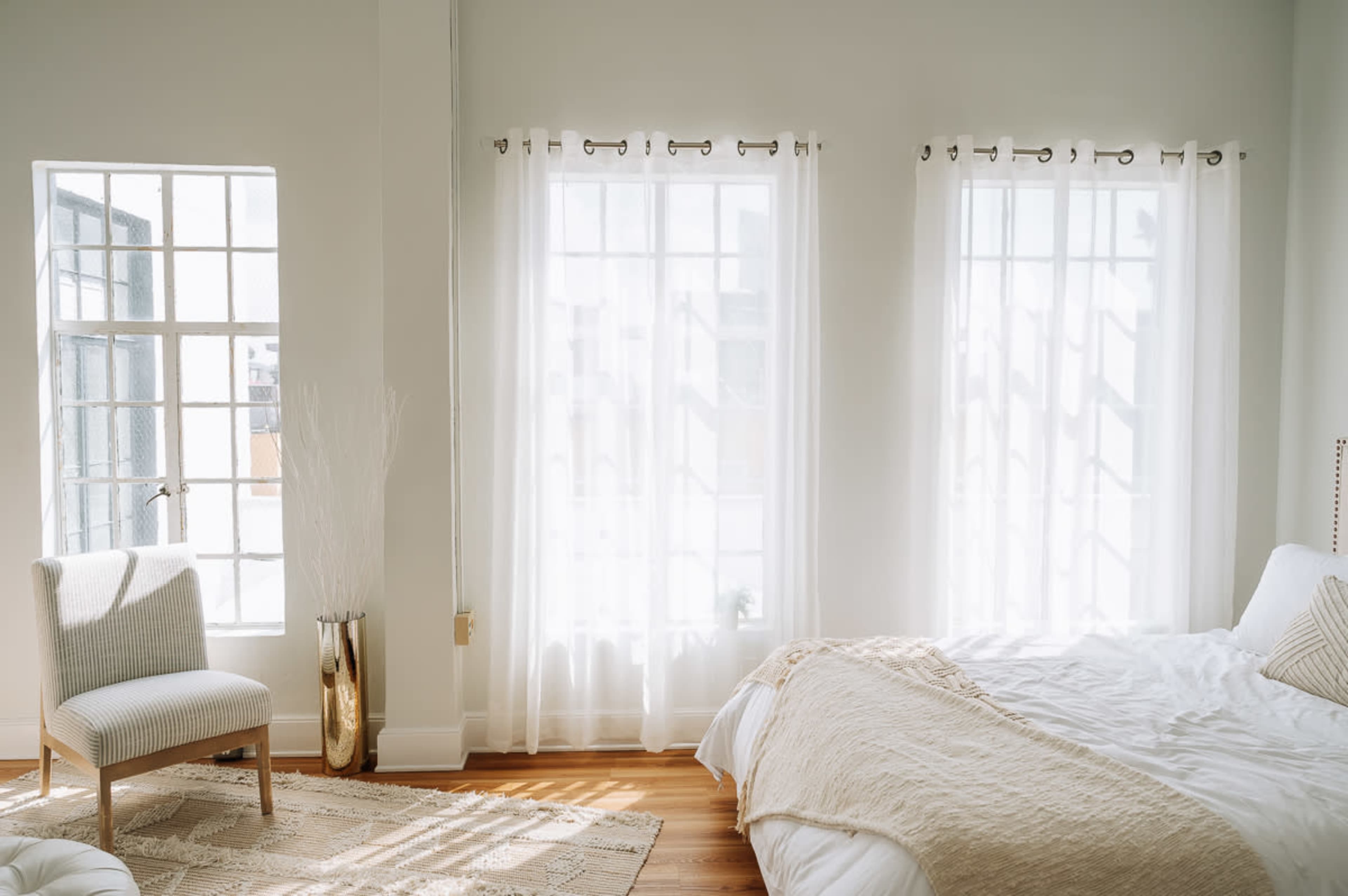The image shows a bright, minimalistic bedroom with large windows adorned with sheer curtains, a bed covered in white linens, and a chair beside a decorative vase.