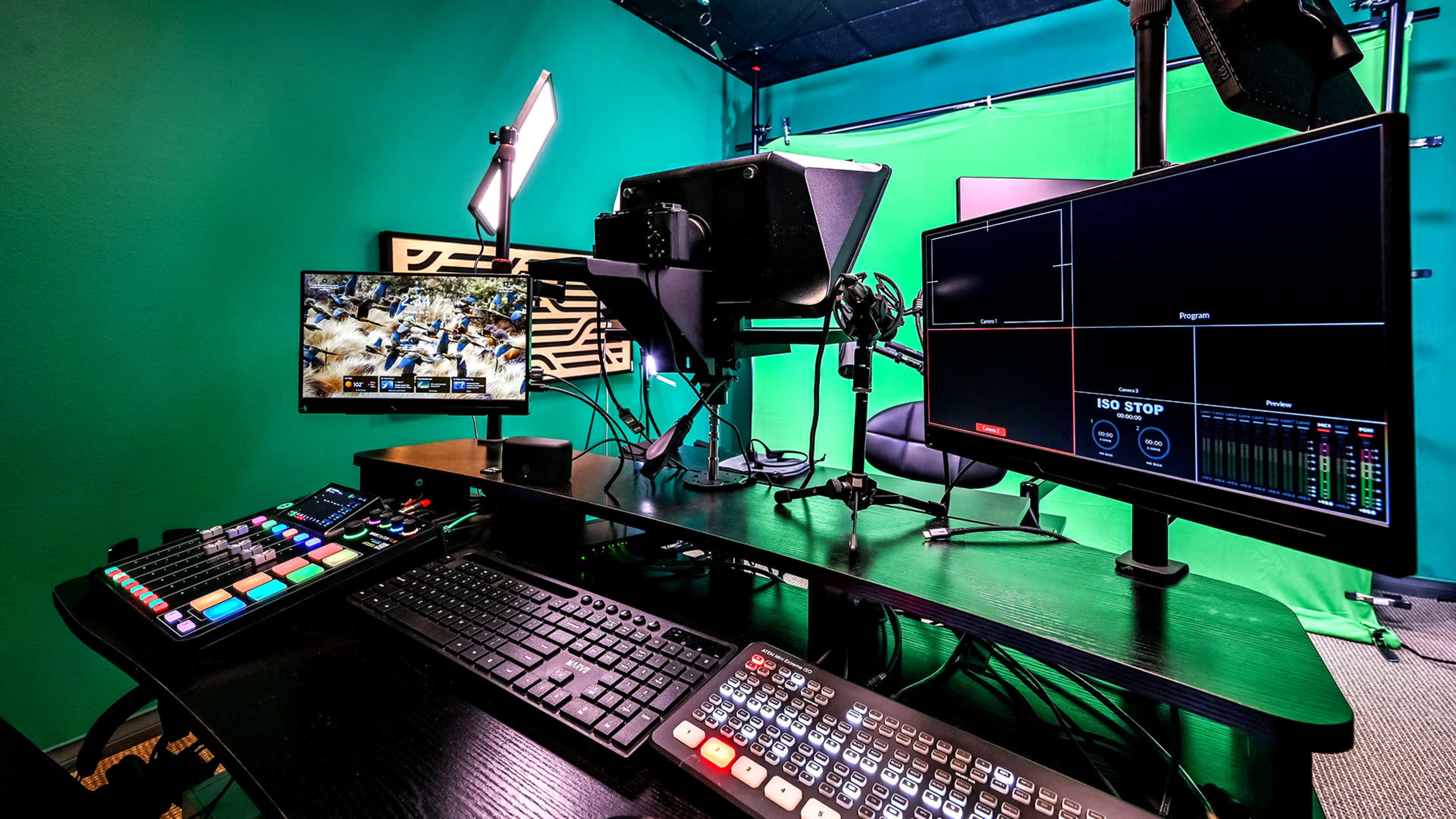 The image shows a video production setup featuring multiple monitors, a keyboard, a control panel, and studio lighting against a green backdrop.
