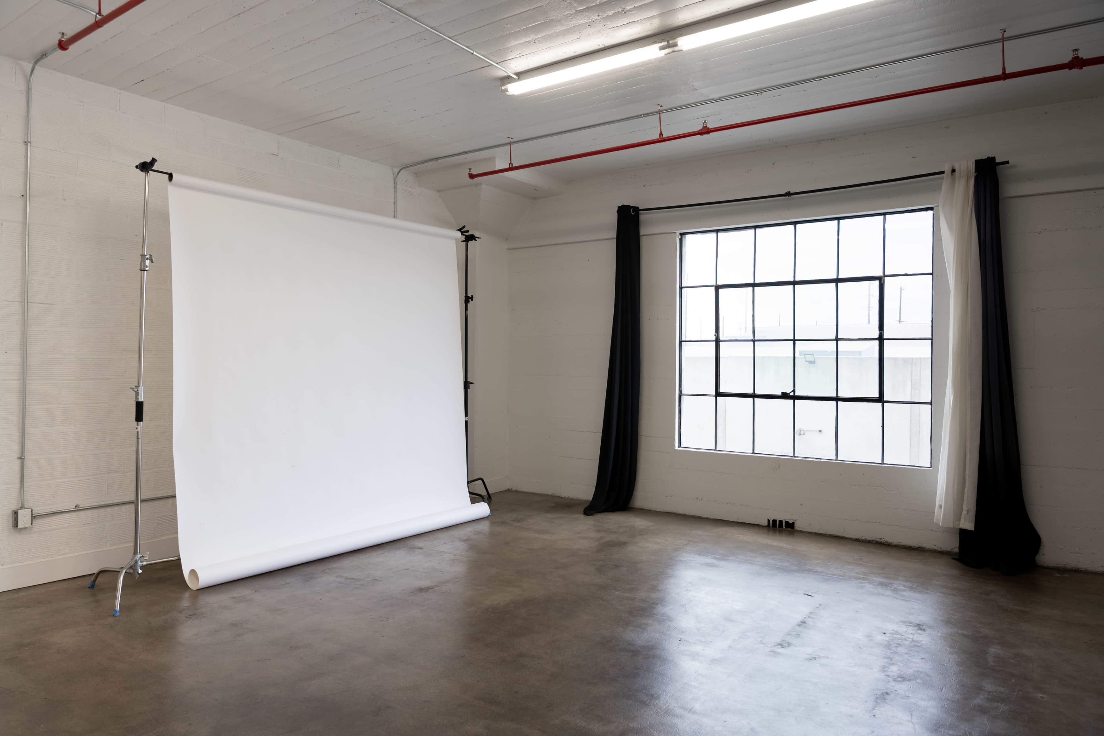The image shows a minimalist studio space with a white backdrop on a support stand and a large window featuring a black and white curtain.