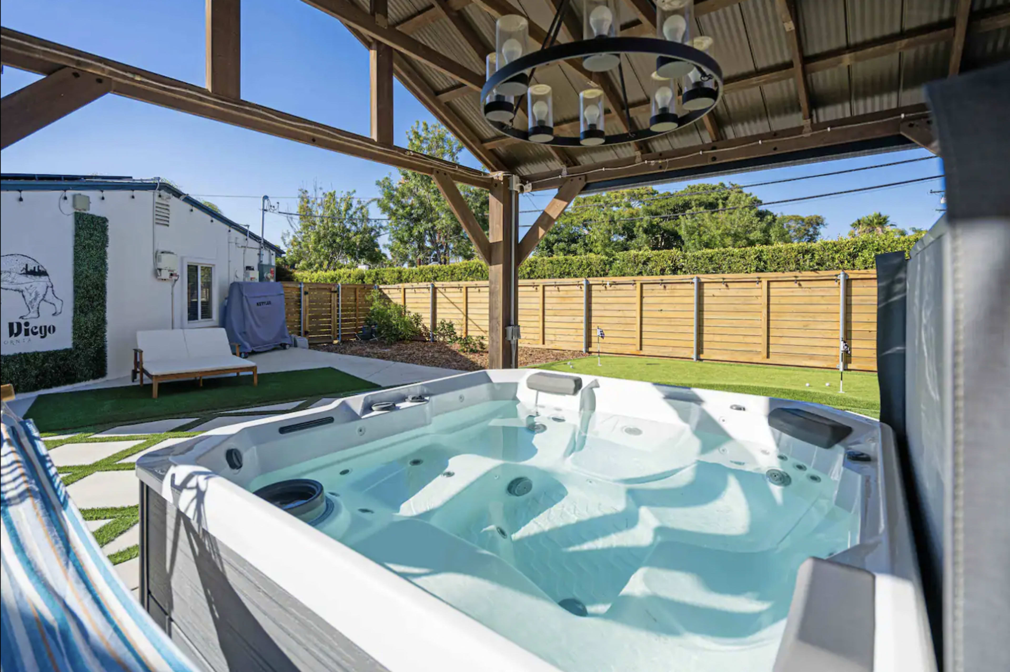 A hot tub is positioned under a wooden gazebo in a backyard surrounded by a wooden fence and landscaped grass.
