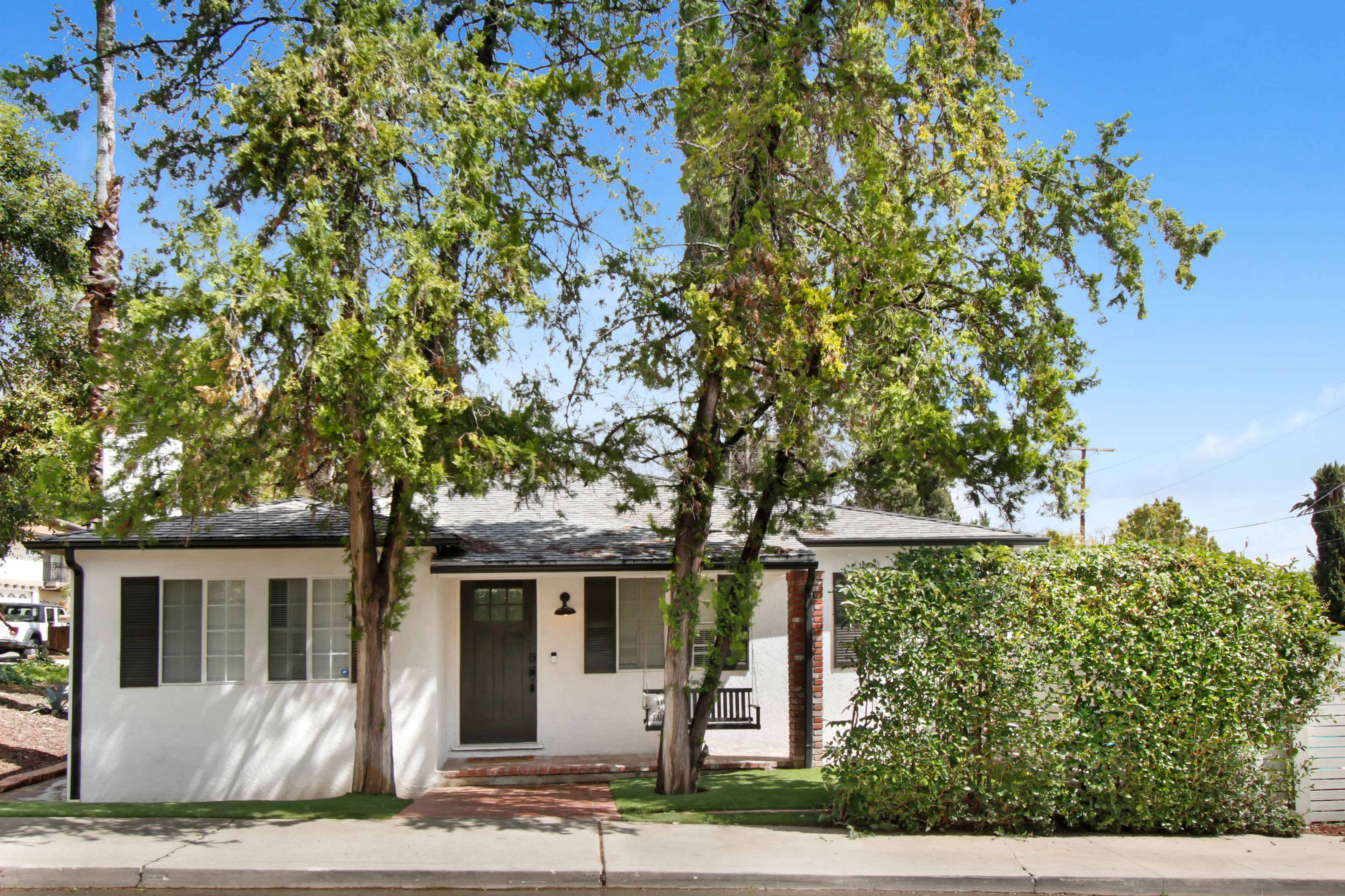 A single-story, white-painted house with green shutters is surrounded by trees and shrubs along a paved walkway.