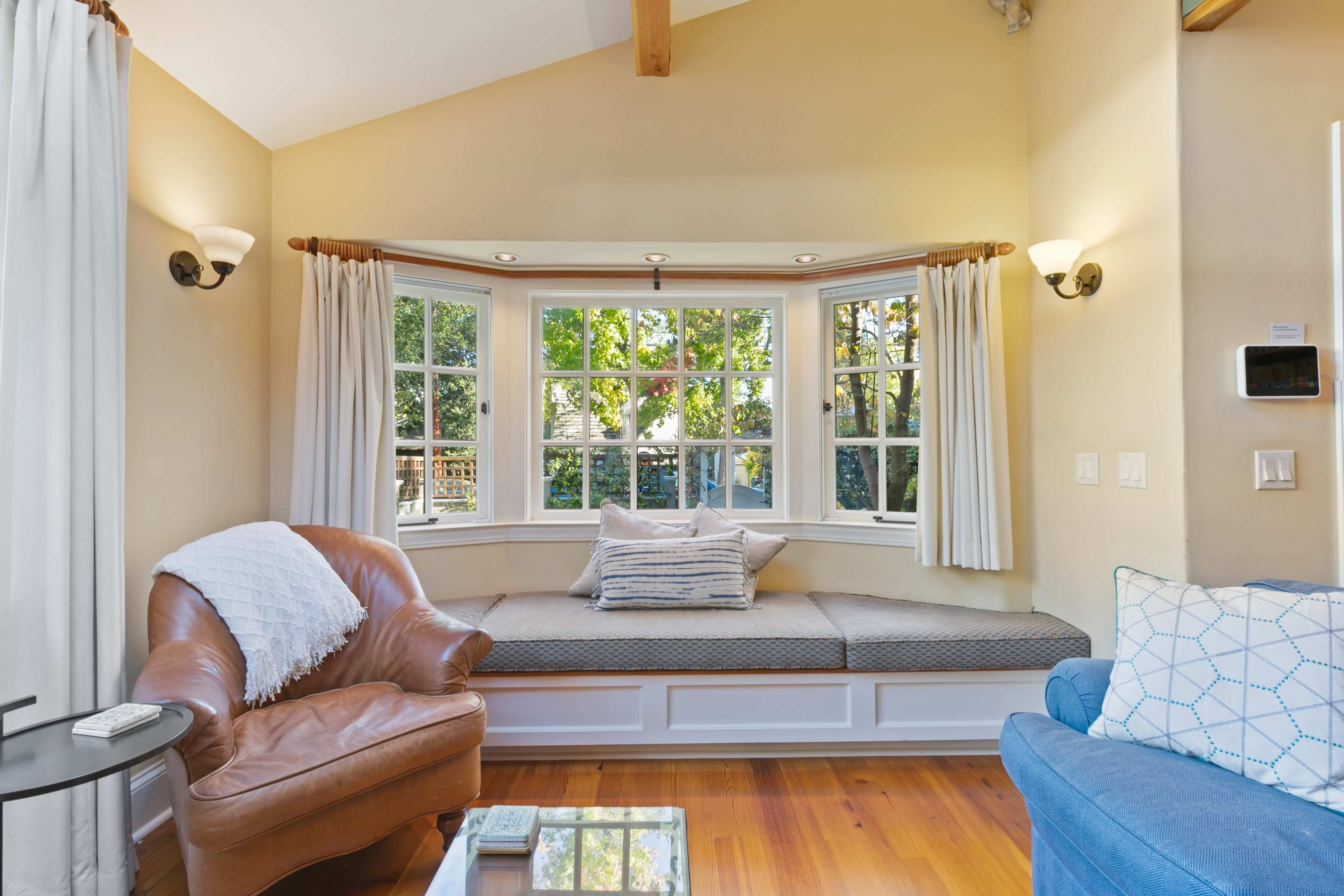 A cozy nook with a cushioned window seat surrounded by large windows, framed by light curtains, and a mix of leather and fabric seating in a well-lit room.