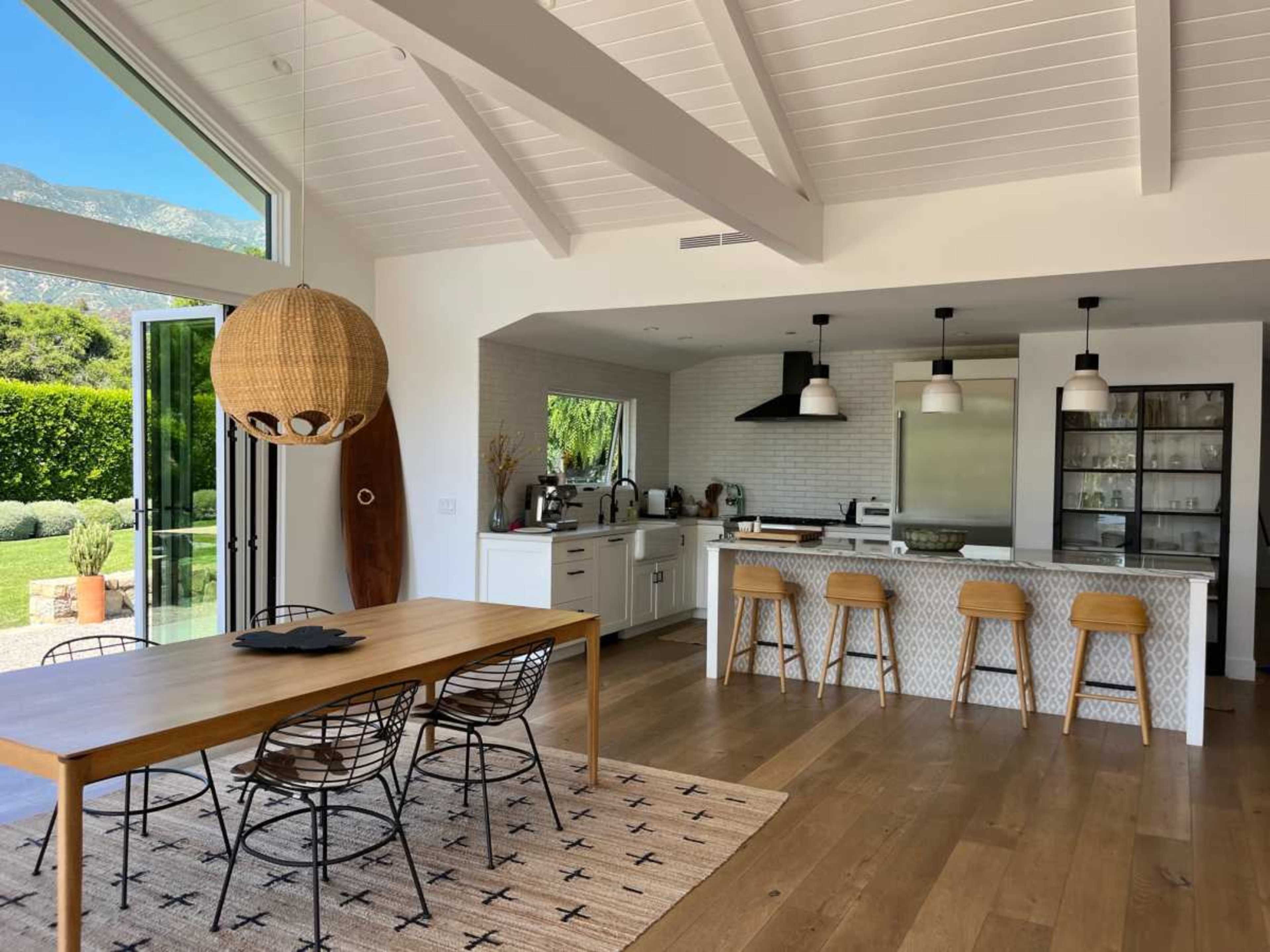 The image shows a modern kitchen and dining area with a wooden table, bar-style seating, and large windows overlooking a green outdoor space.