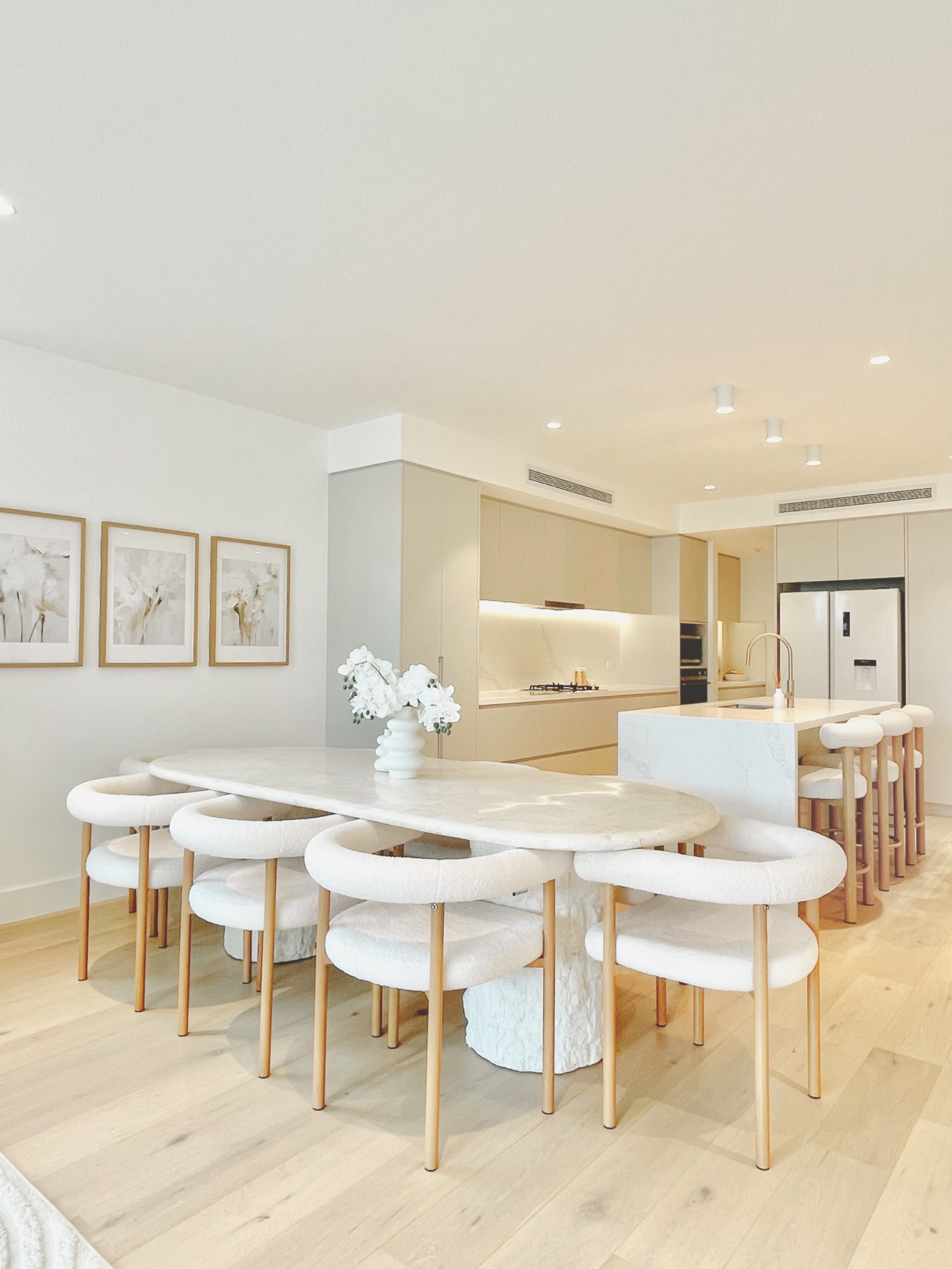 The image shows a modern kitchen with a large marble dining table surrounded by circular white chairs, framed artwork on the wall, and a minimalist design.