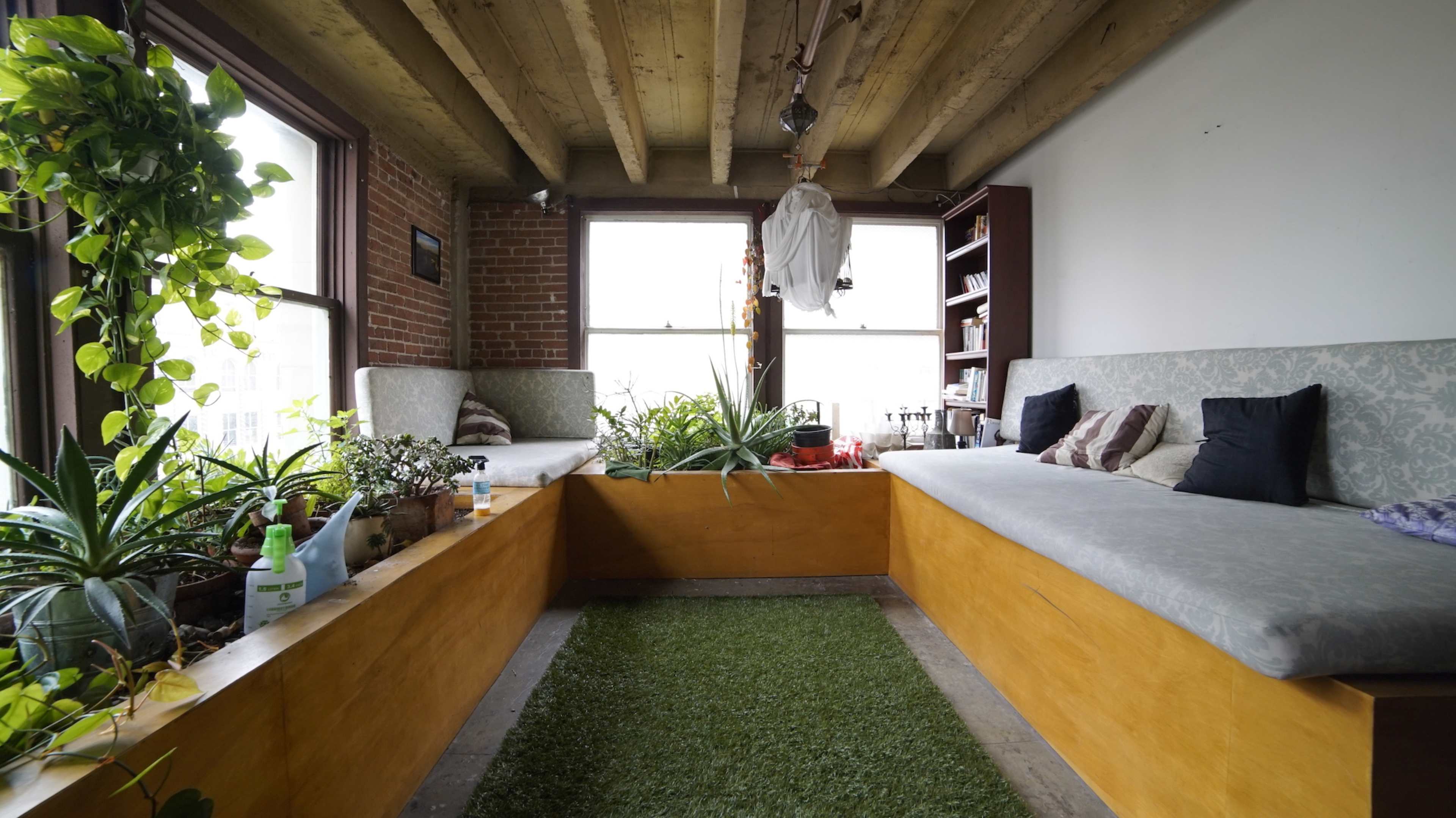 The image shows a cozy room with two cushioned benches surrounding a patch of artificial grass and various plants lining the windows.