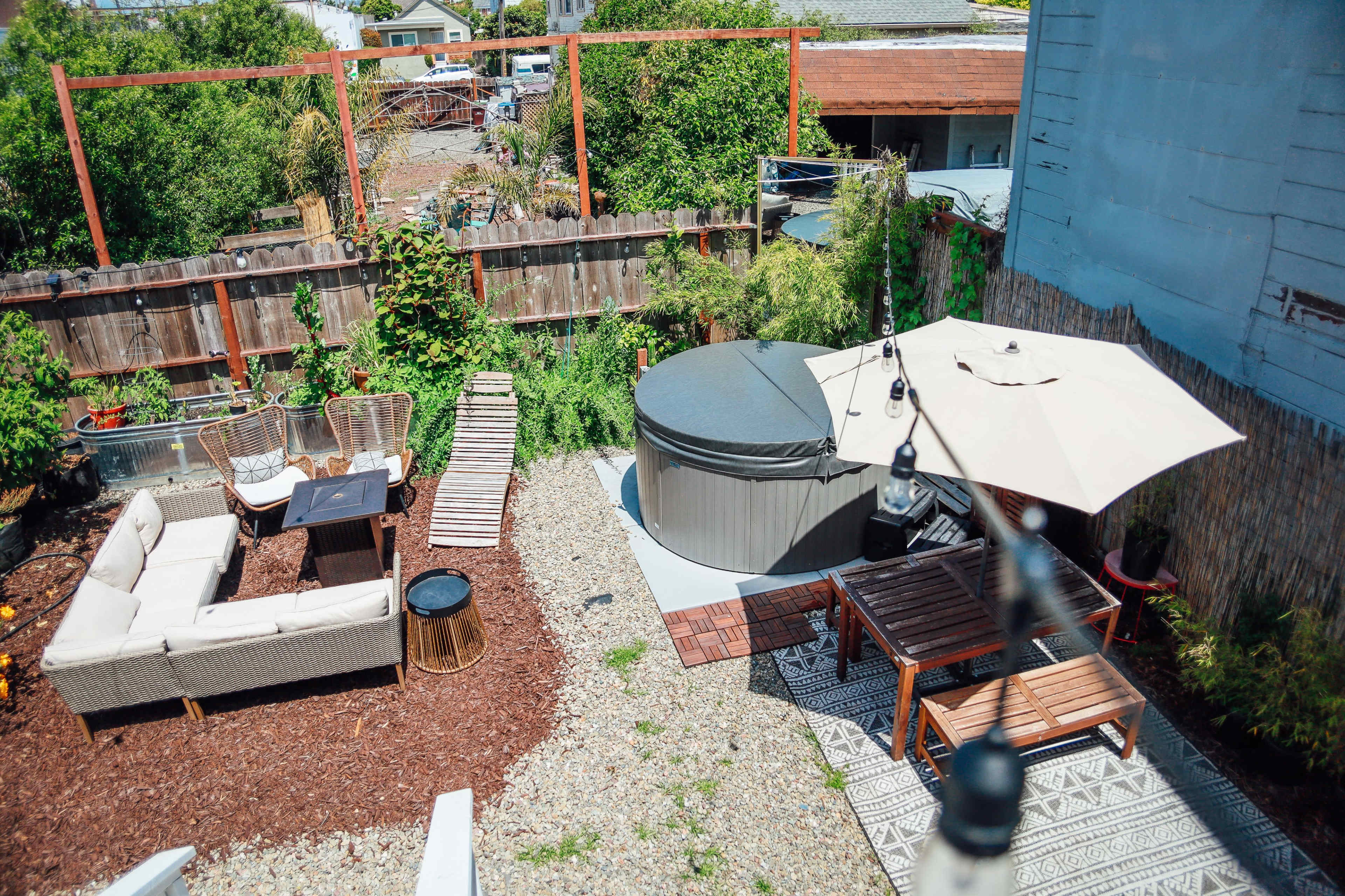 A landscaped backyard with a seating area, hot tub, and a wooden dining table under a large umbrella.