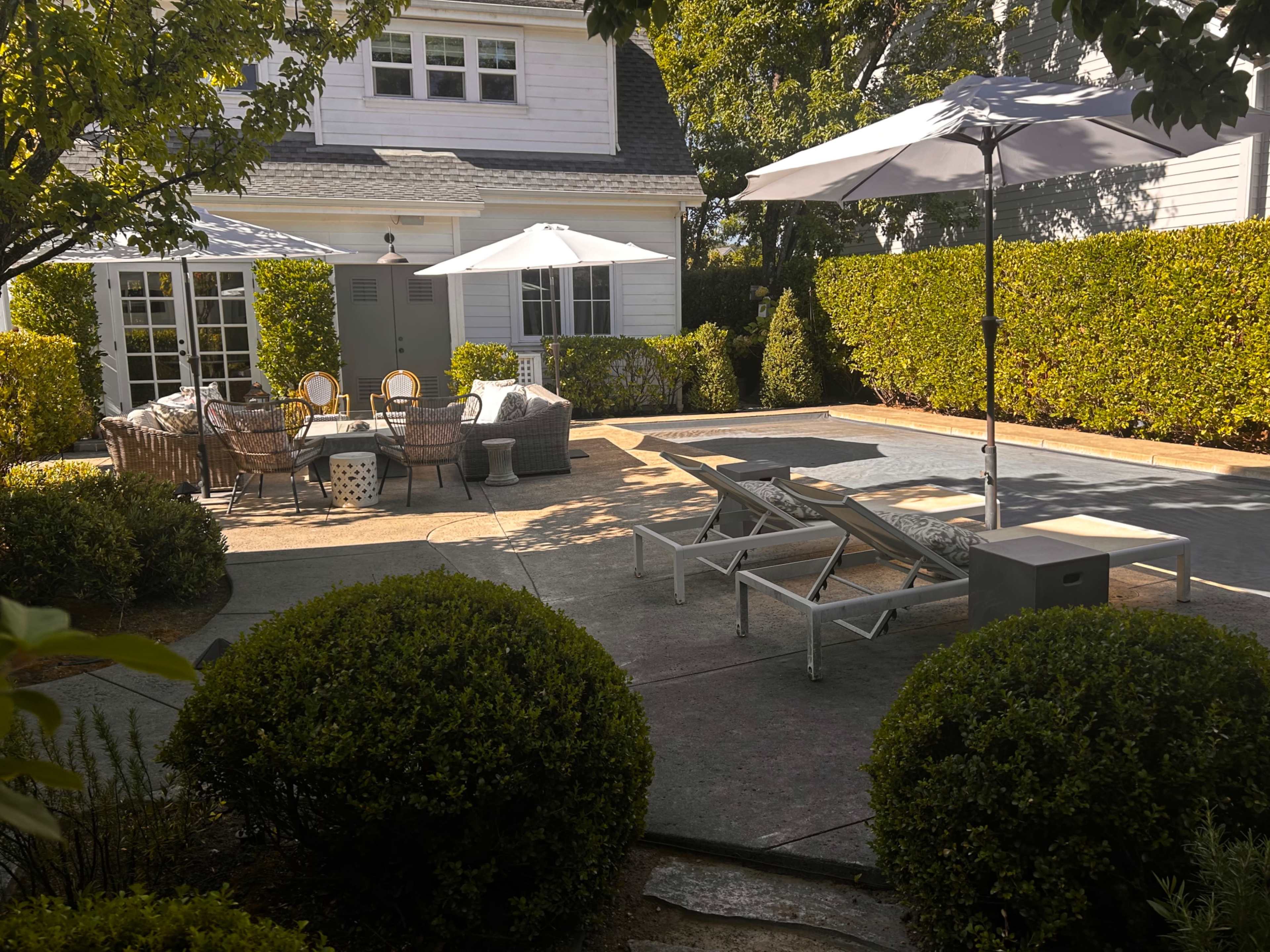 The image shows a well-arranged outdoor patio area with seating, umbrellas, and lounge chairs surrounded by shrubs and trees.