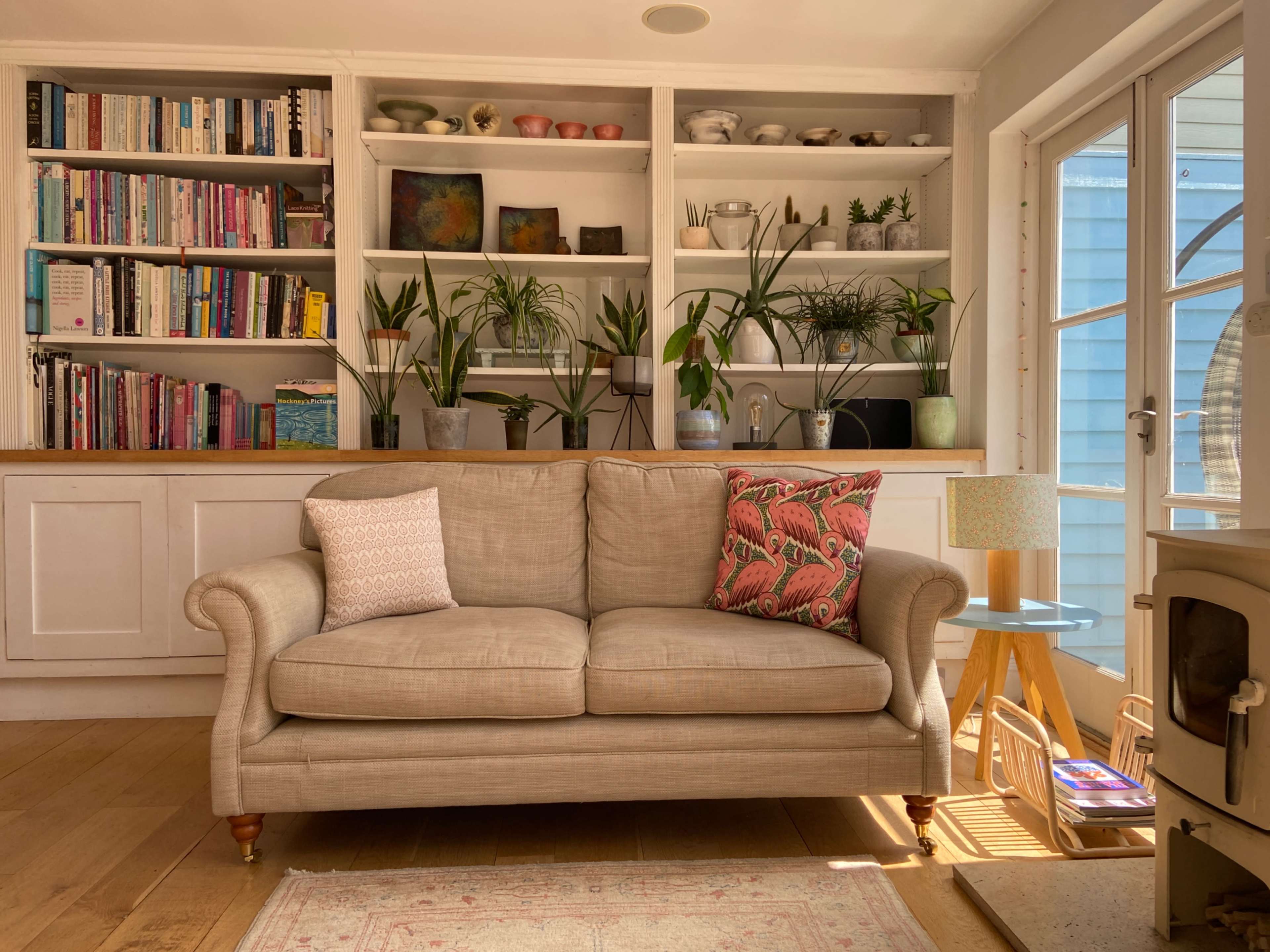 A beige sofa with decorative cushions is positioned in front of a bookshelf filled with books and plants, alongside a small side table and a warm light source.