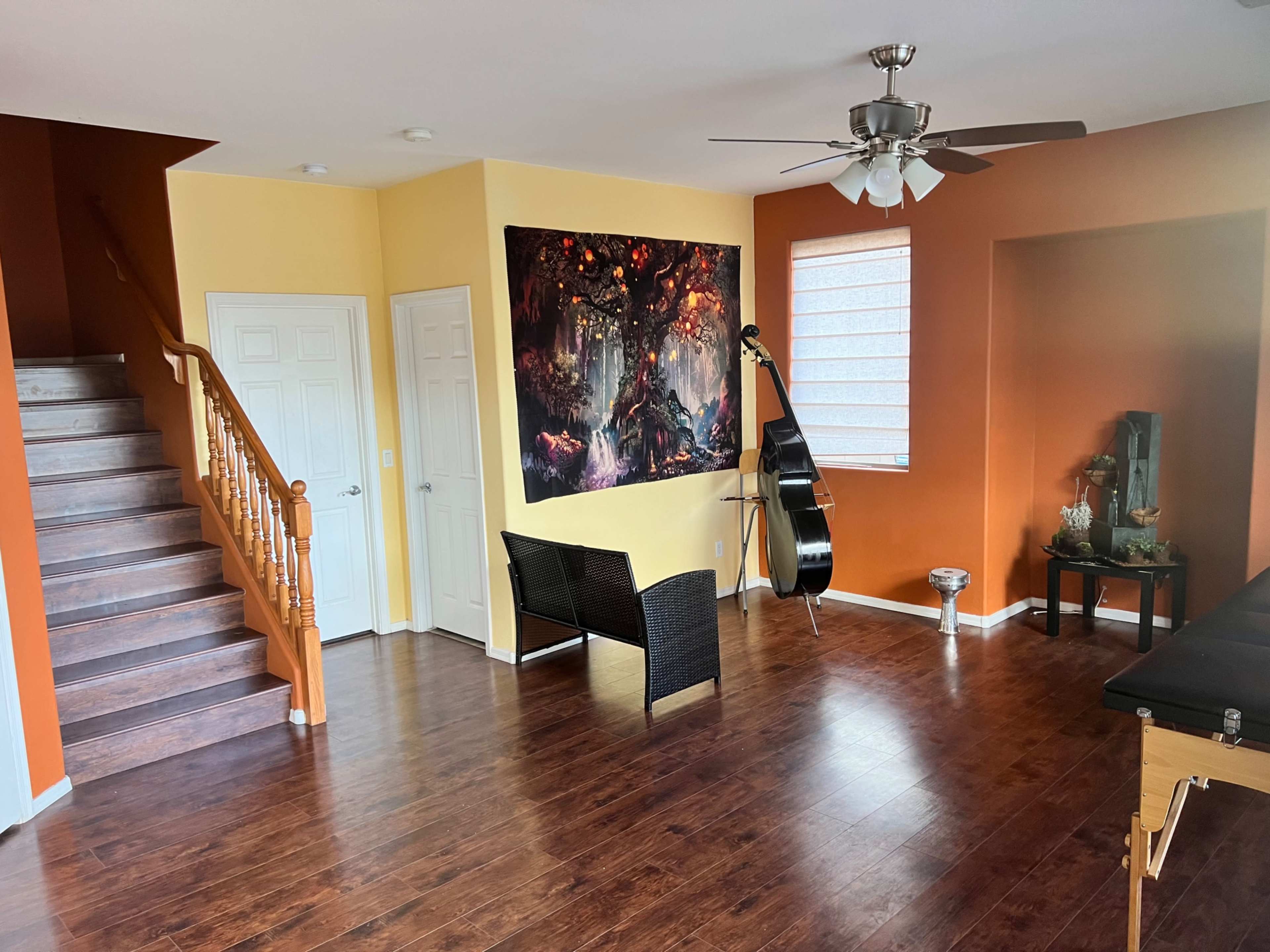 A spacious living area with wooden flooring, a staircase on the left, a large colorful painting on the wall, and a few pieces of furniture including a black chair and a tall, narrow mirror.