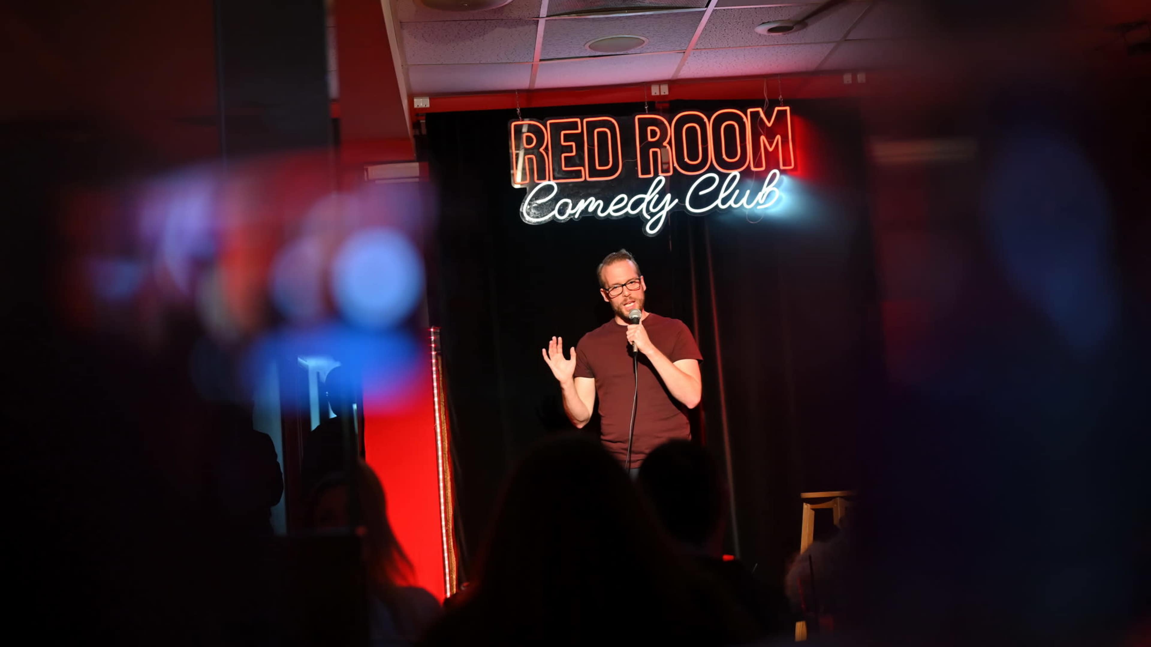A comedian performs on stage at the Red Room Comedy Club, illuminated by a neon sign.