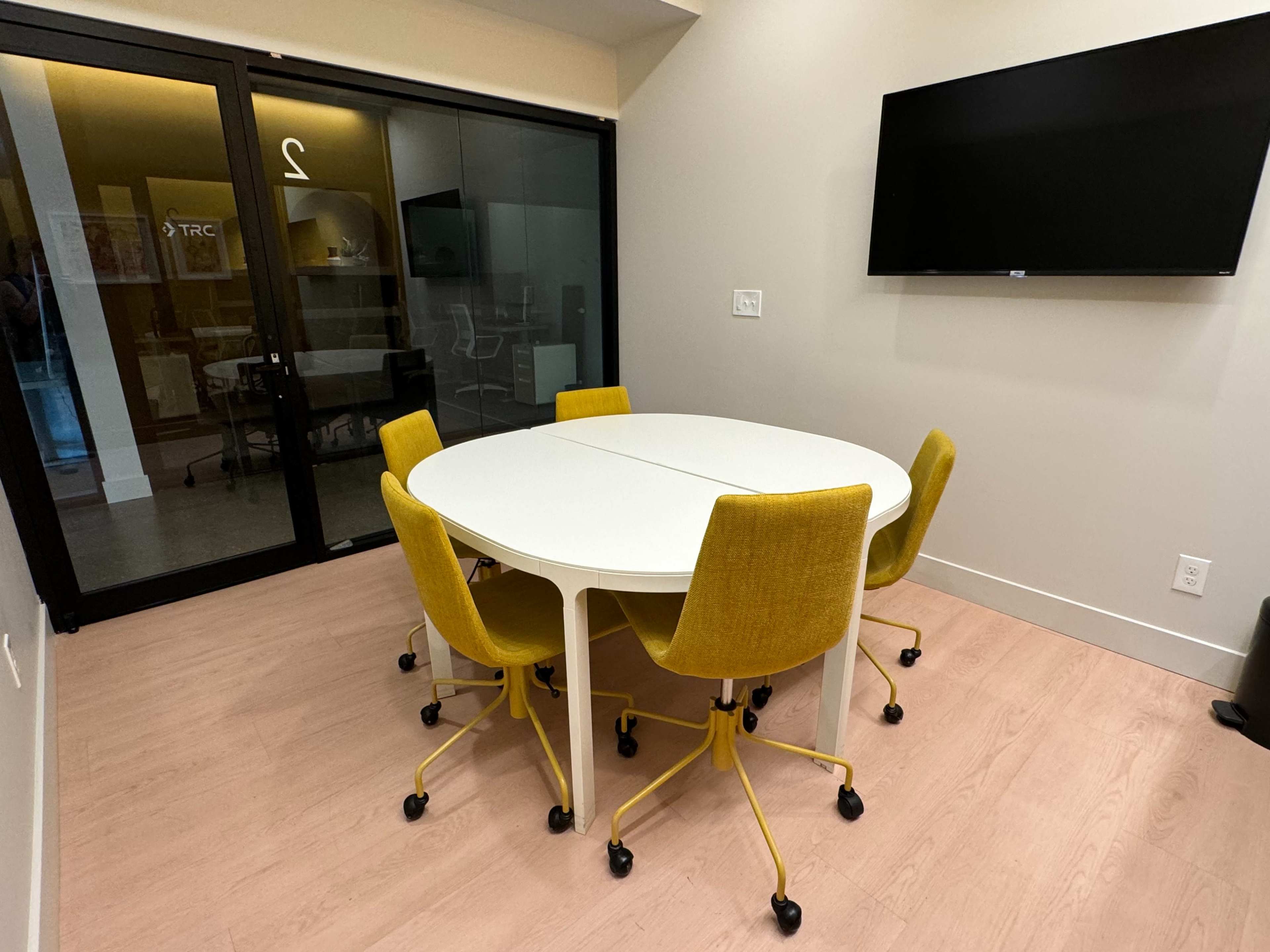 A round white table with four yellow chairs is set up in a conference room with a large screen and glass doors.