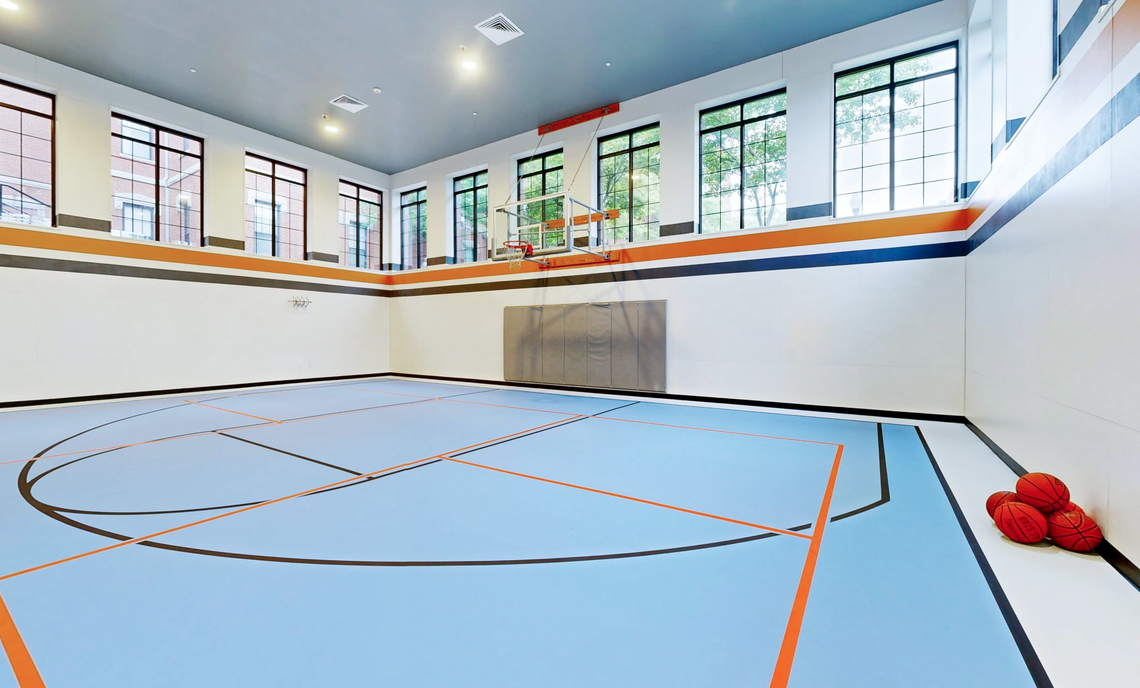 The image shows a well-lit indoor basketball court with a blue floor, orange lines, and two basketballs resting in the corner.