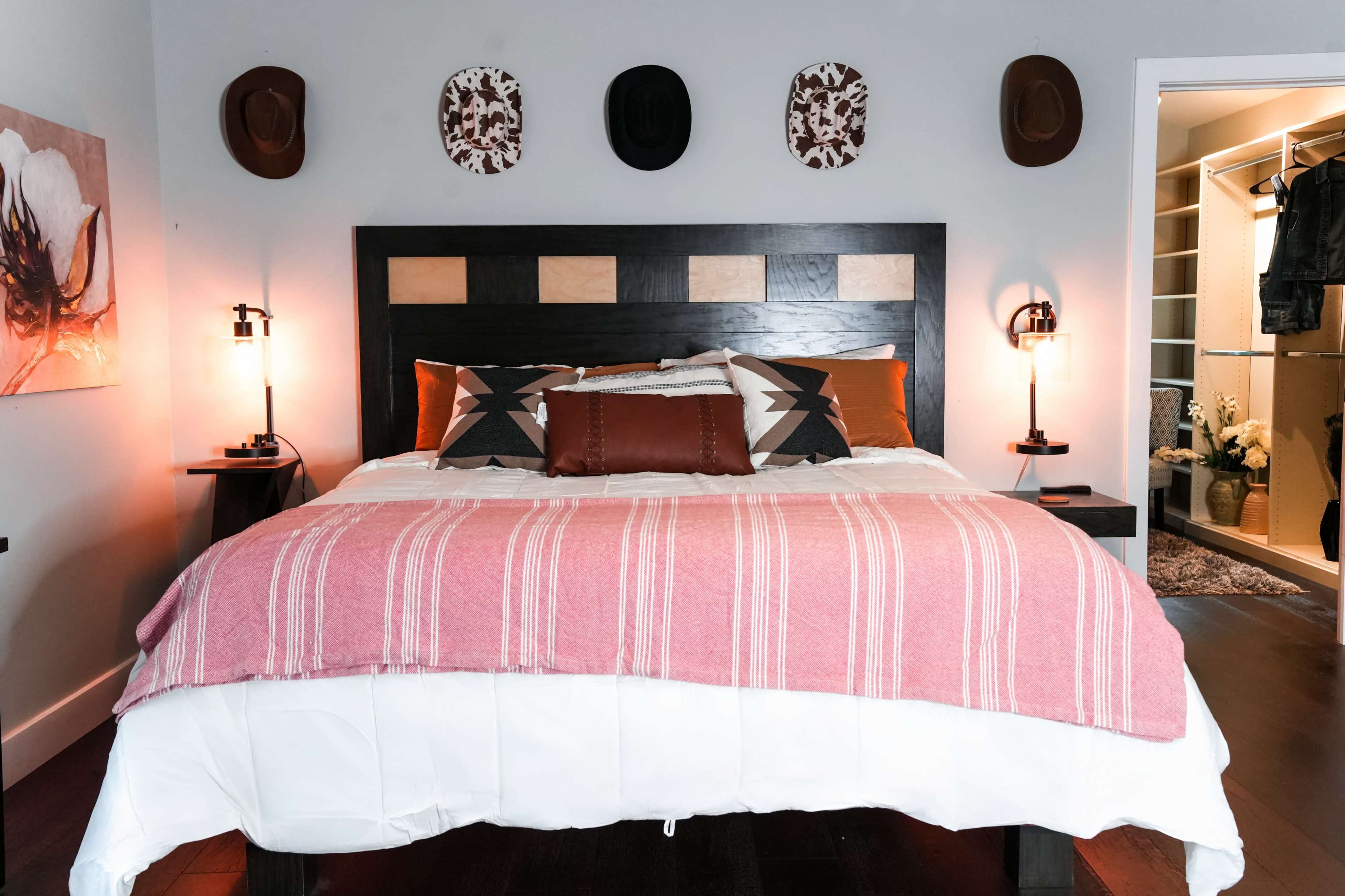 The image shows a well-decorated bedroom featuring a large bed with a pink and white striped blanket, two bedside lamps, and decorative hats mounted on the wall above the bed.