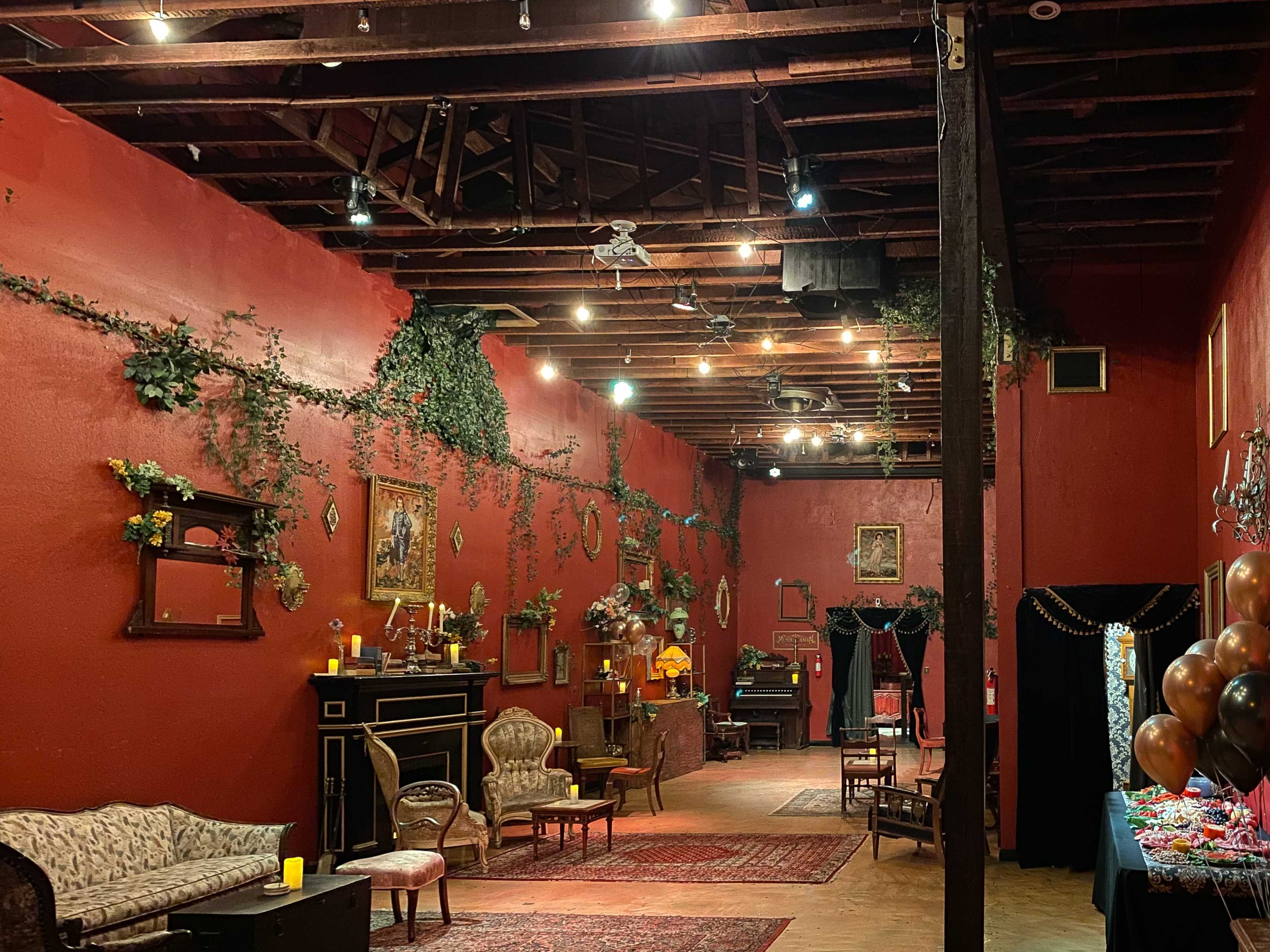 The interior of a dimly lit venue features red walls adorned with greenery, vintage furniture, and a small stage area with a piano.