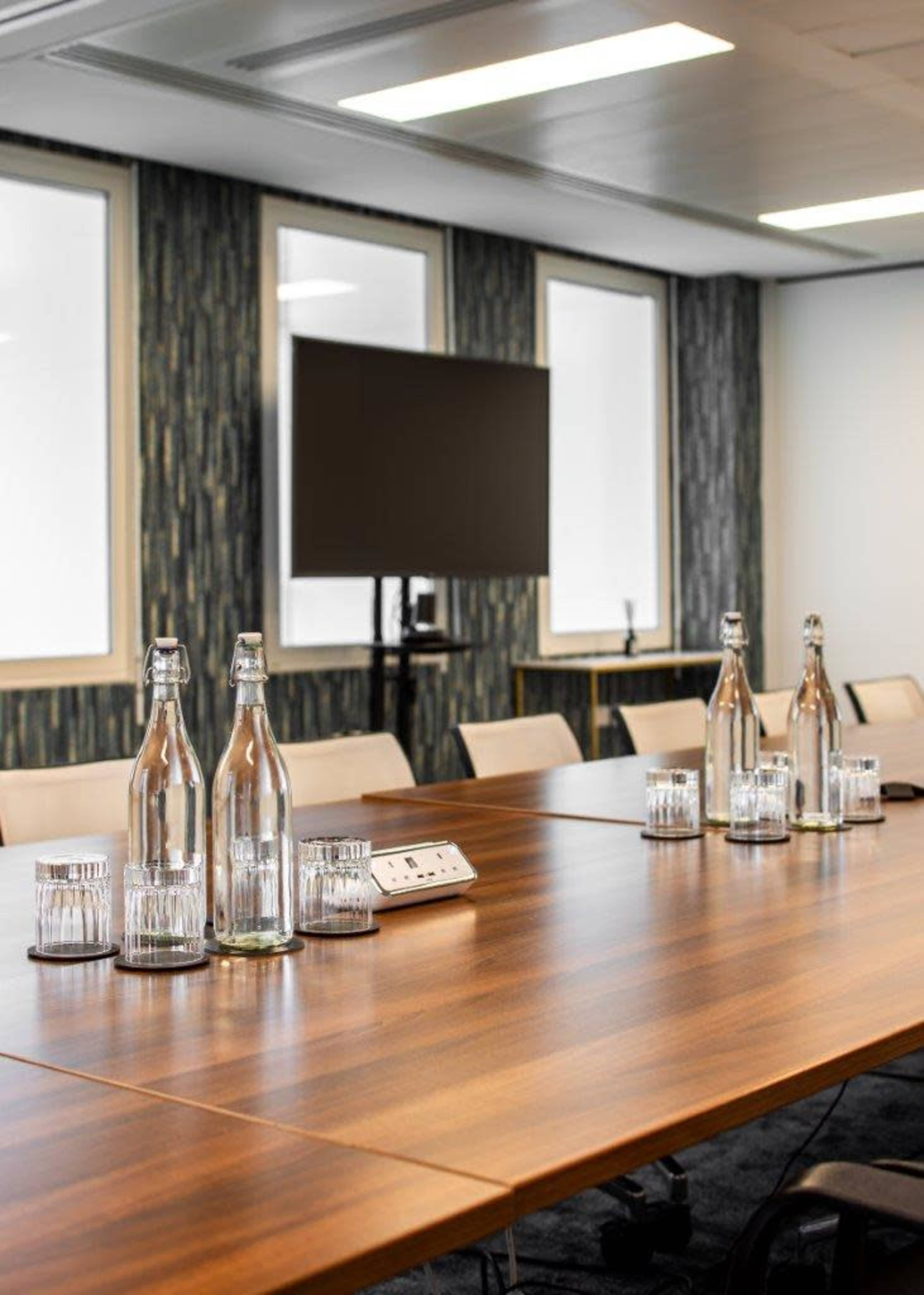 A modern conference room features a wooden table set with glass water bottles and short glasses, accompanied by a wall-mounted display screen.