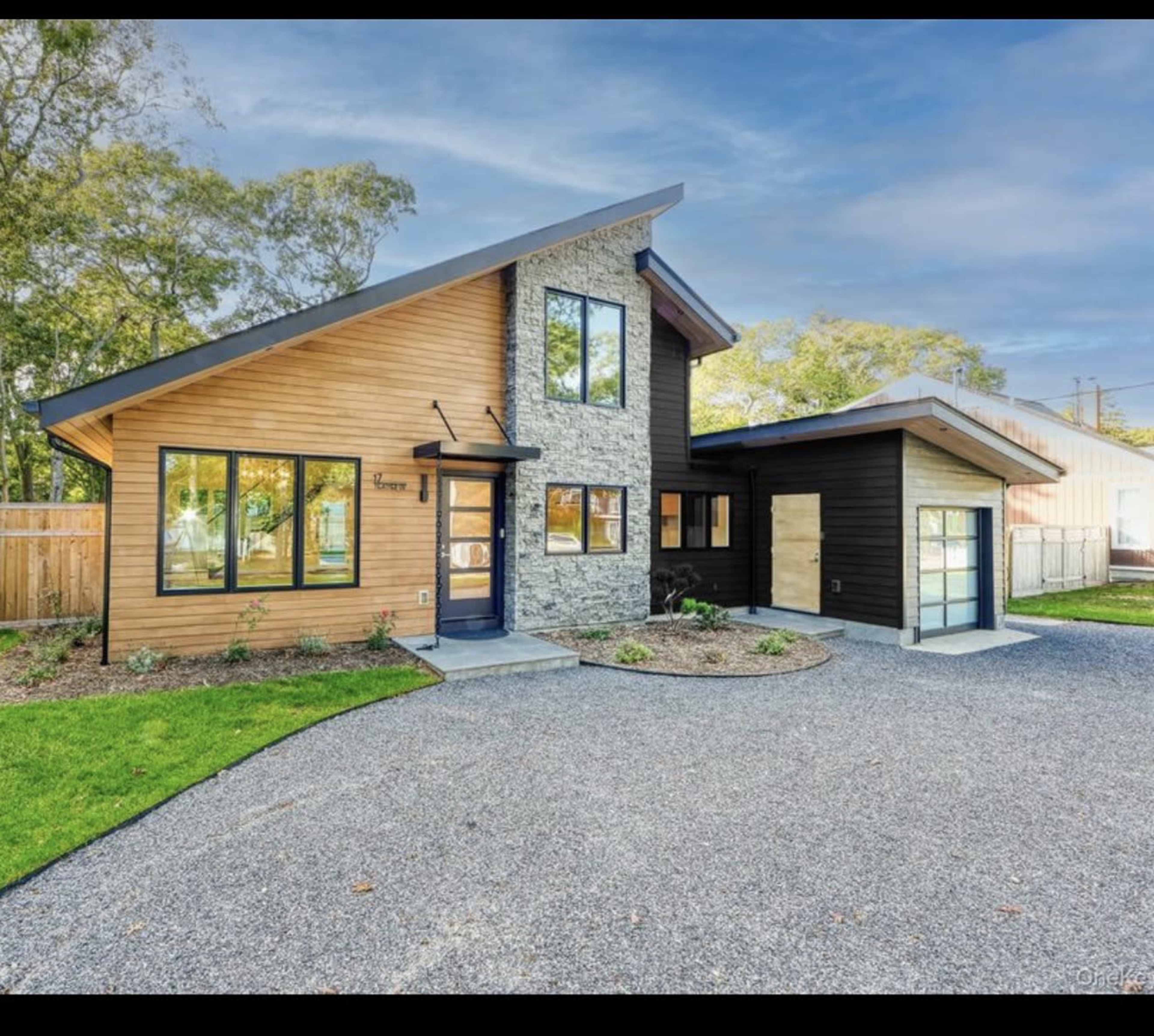 The image shows a modern two-story house with a combination of wood and stone siding, a unique angled roof, and a circular gravel driveway.