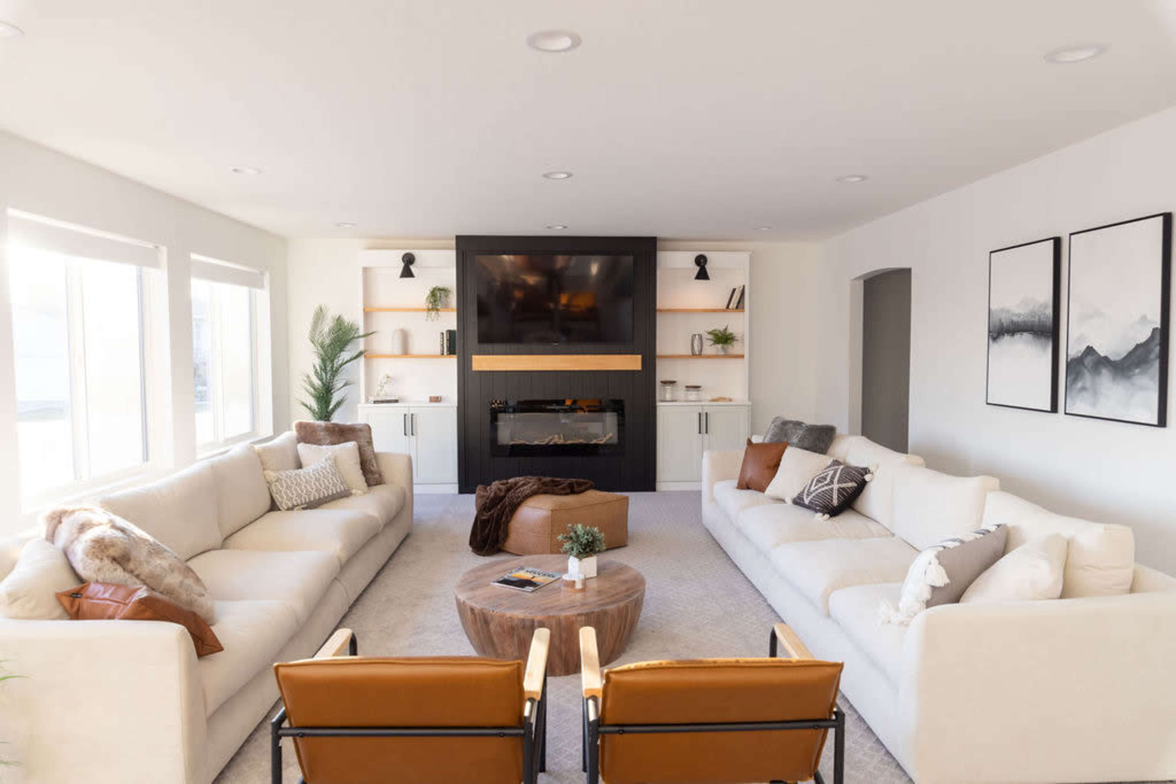 A modern living room with two symmetrical sofas, two wooden armchairs, a round coffee table, and a wall-mounted television above an electric fireplace.