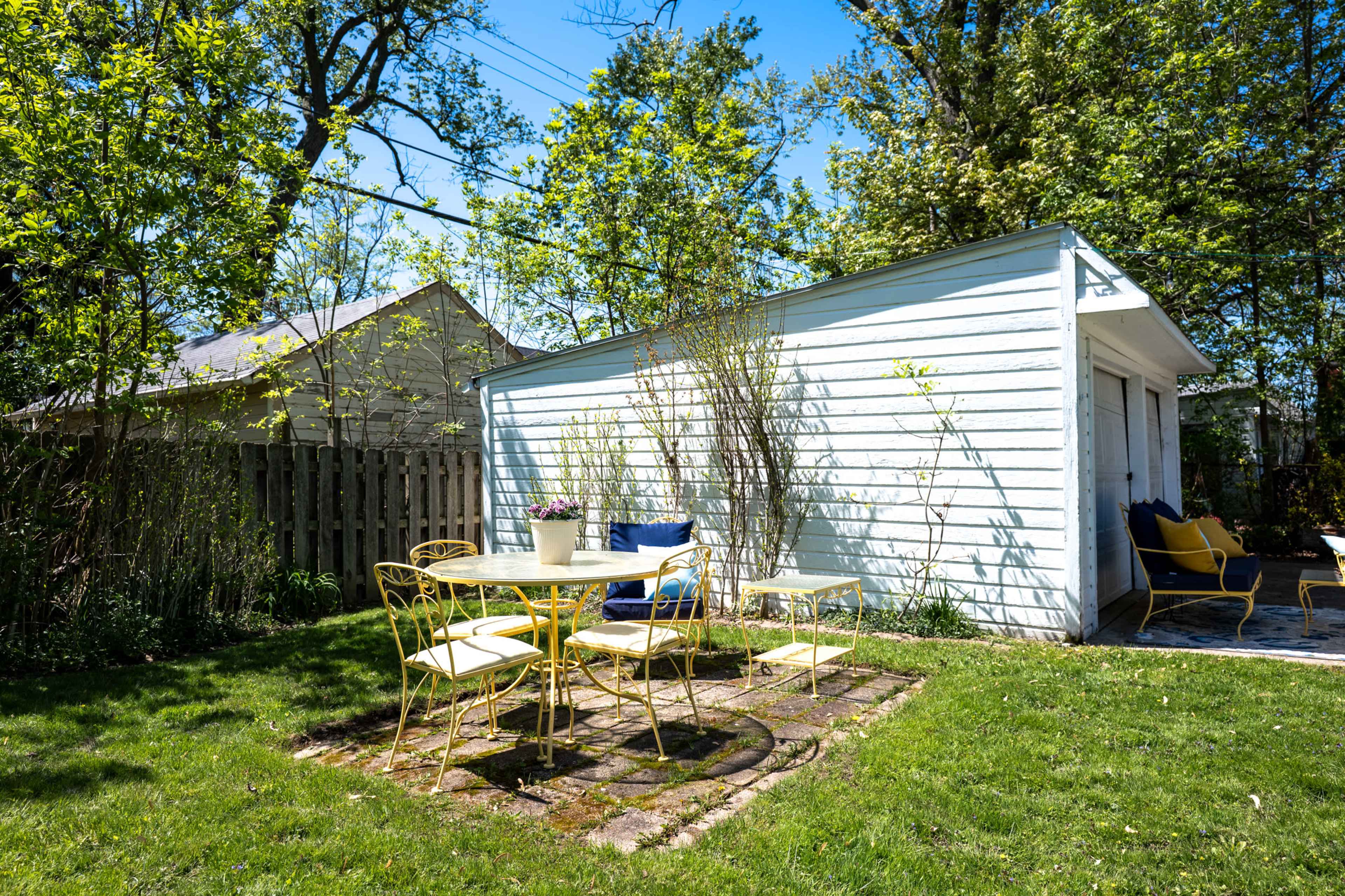 The image shows a white shed surrounded by greenery and a circular yellow metal table with chairs on a grassy area.