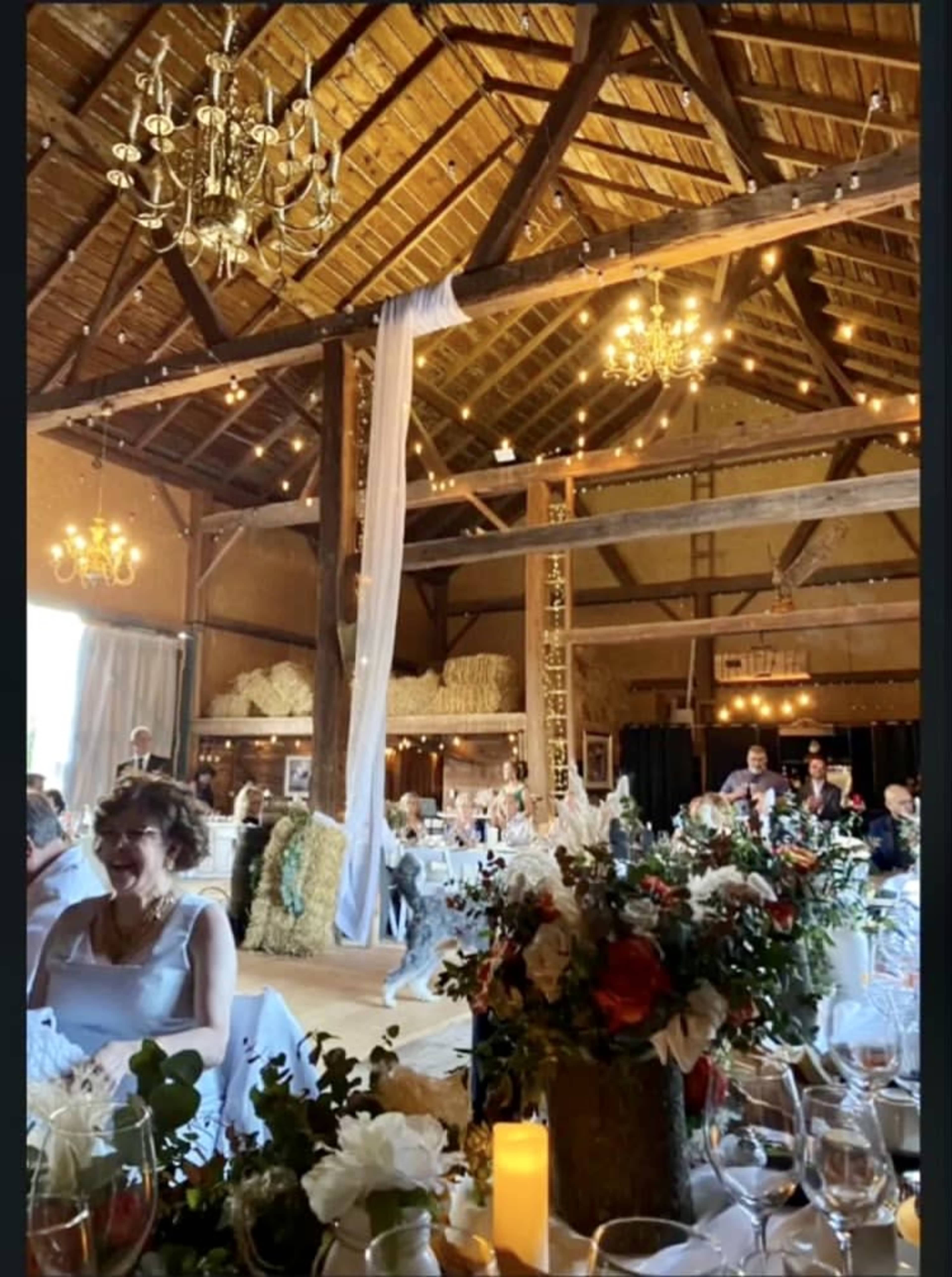 A rustic barn interior decorated for a wedding reception, featuring tables with floral arrangements and soft lighting from chandeliers.