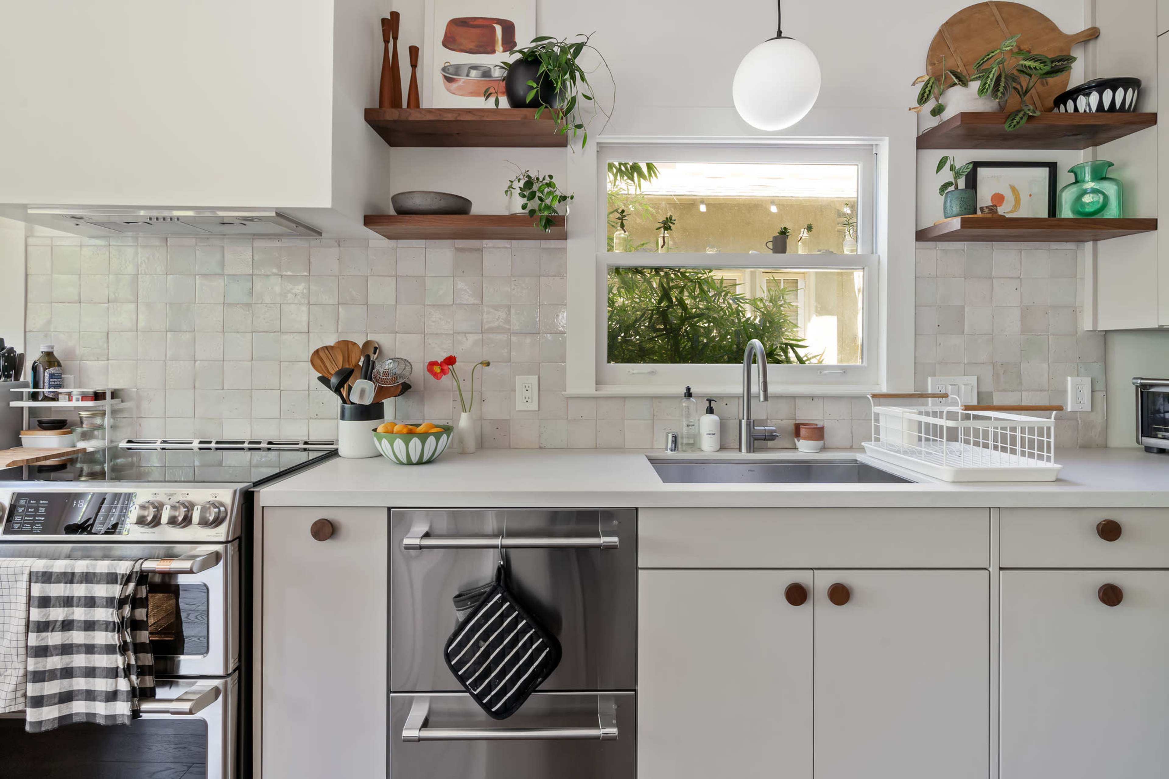 A modern kitchen features a stainless steel sink, open shelves with decorative items, and a window showing greenery outside.