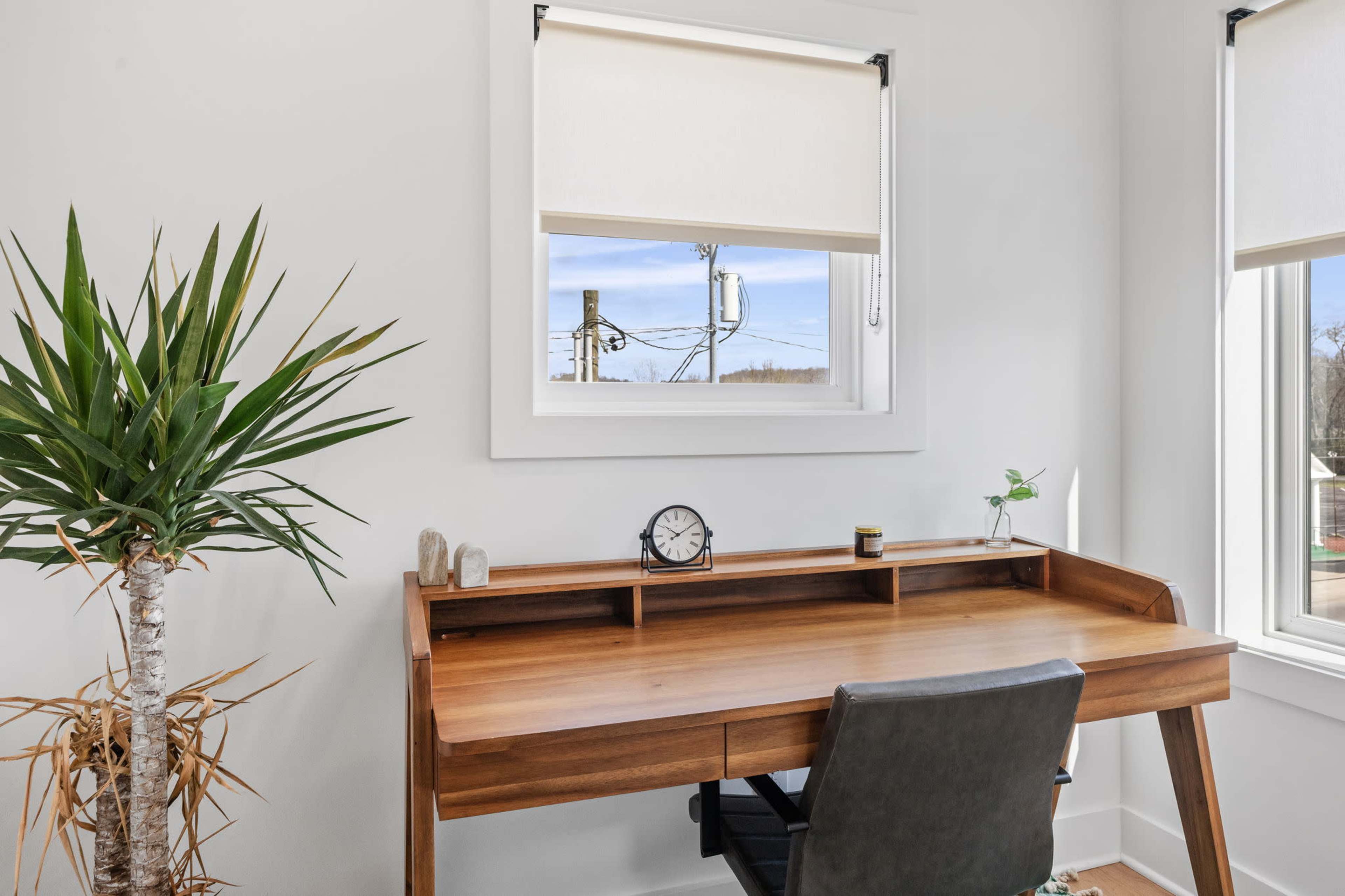 A wooden desk with a clock and a small plant sits by a window that overlooks utility poles and blue sky.