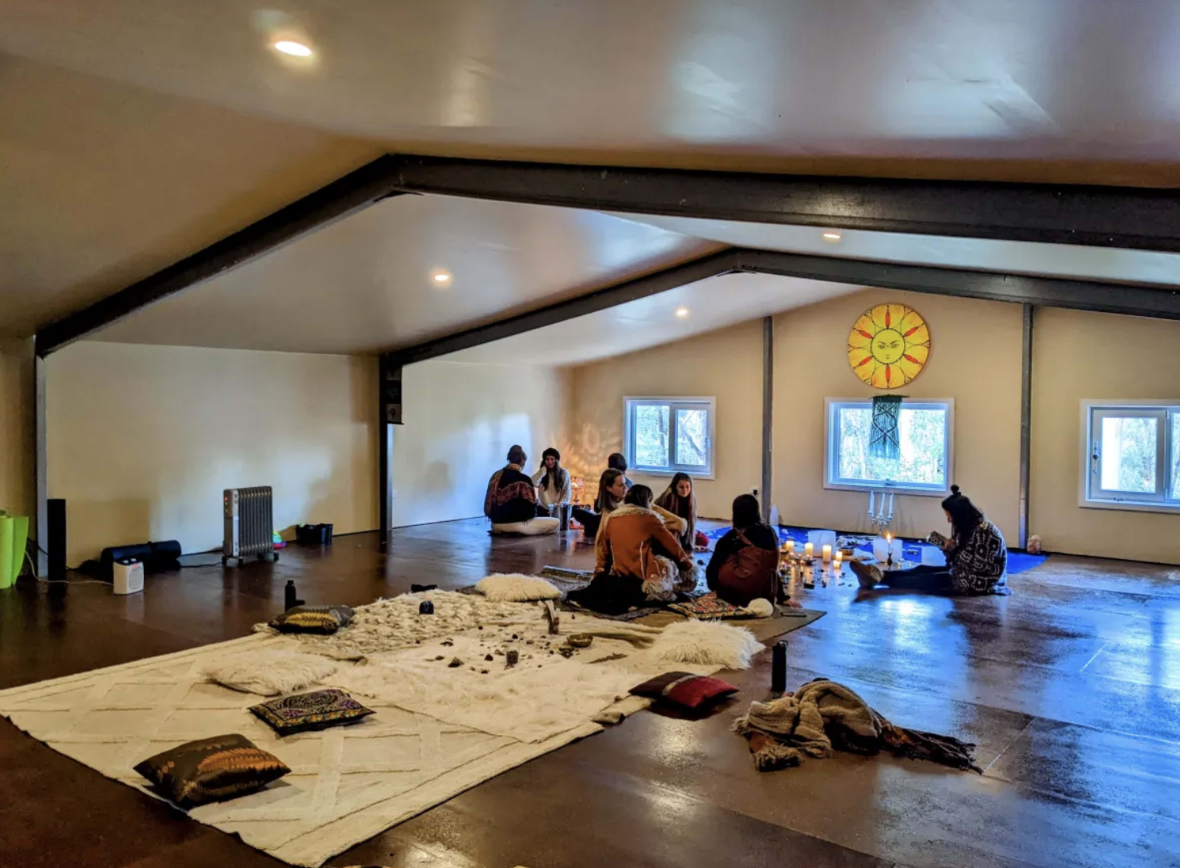 A spacious, light-filled room with people seated on the floor, engaged in an activity near large windows and decorated with cushions and a sun motif on the wall.