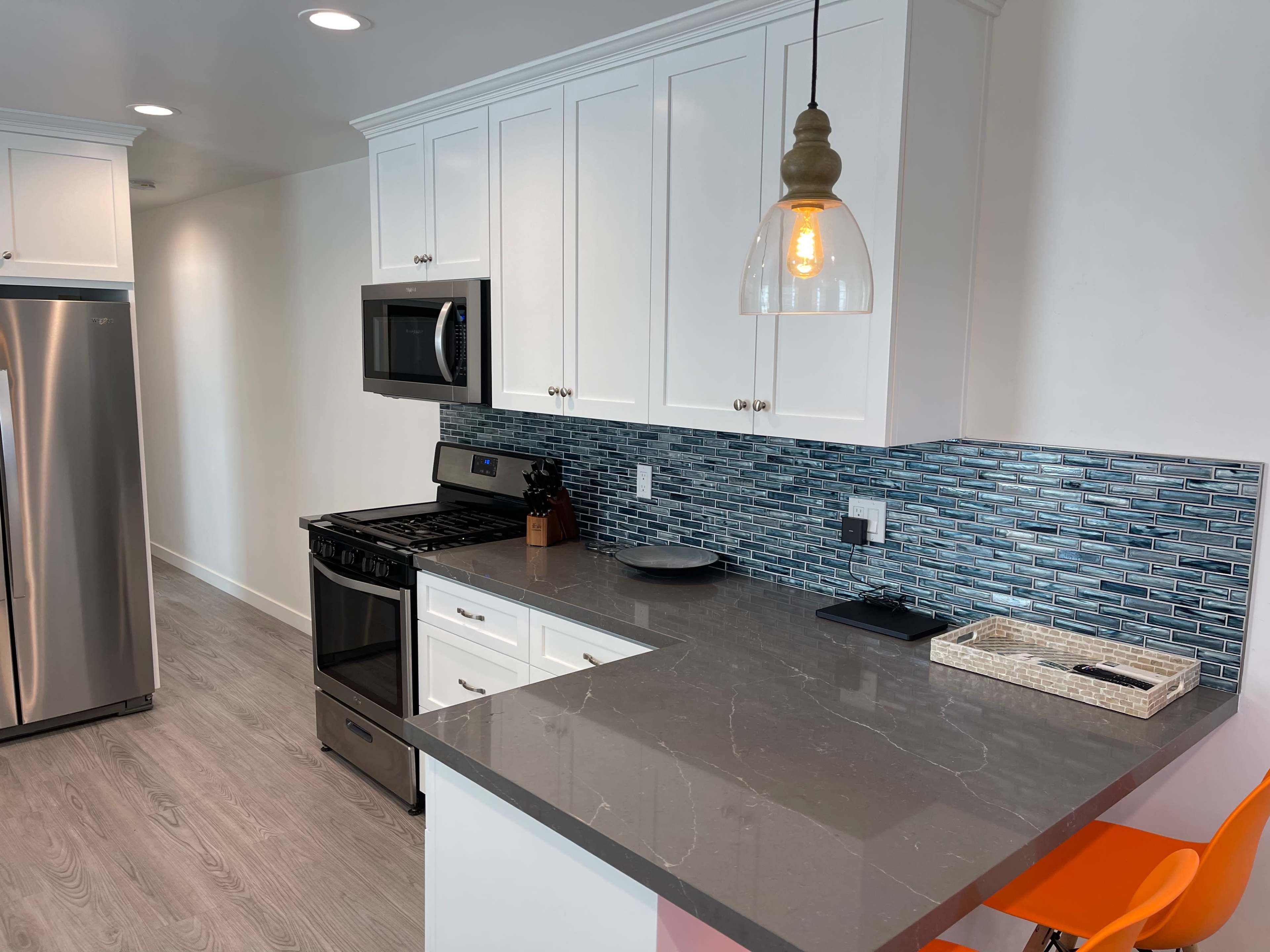 The image shows a modern kitchen with white cabinetry, a gray countertop, stainless steel appliances, and blue tile backsplash.